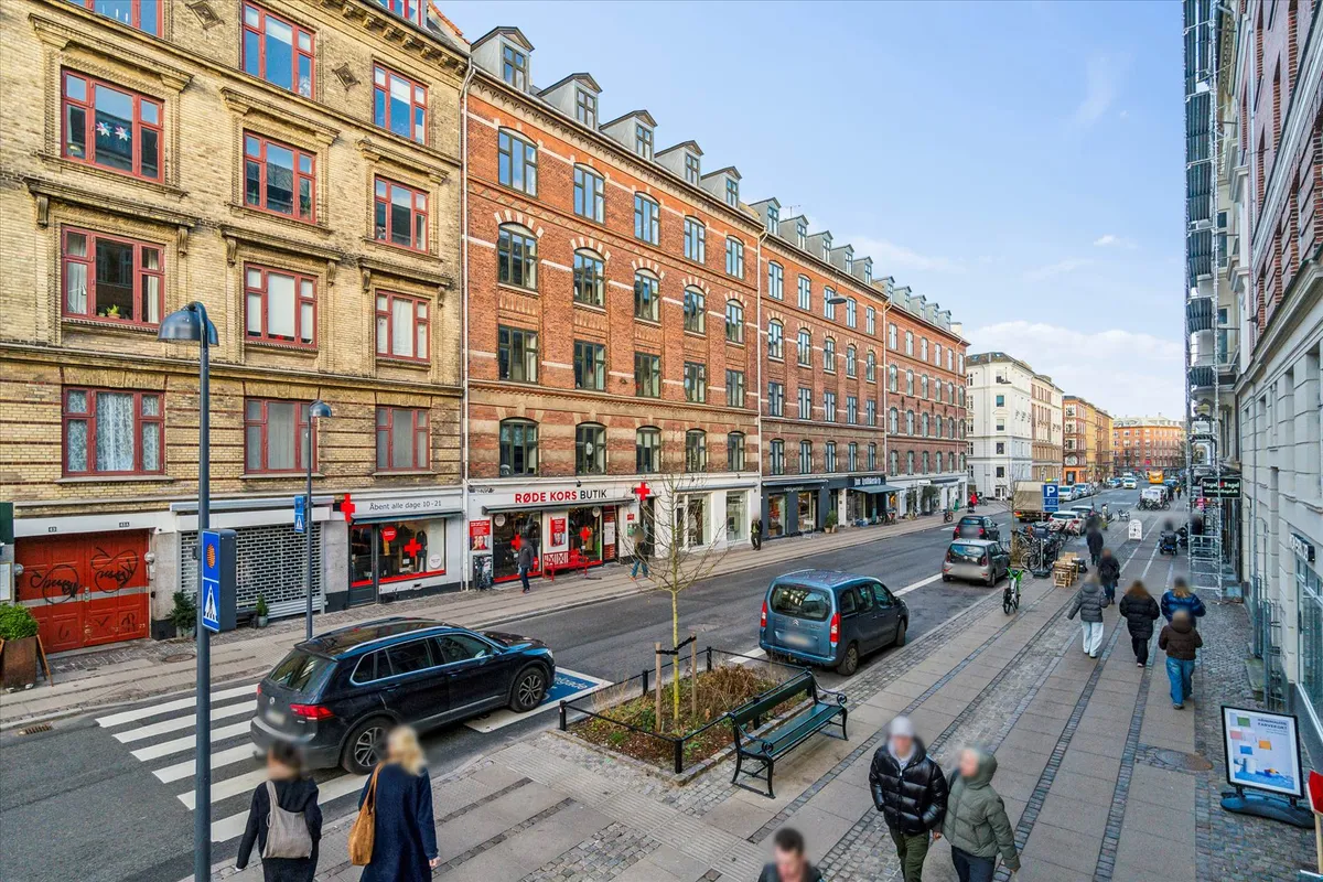 Mennesker går på gågaden med flere biler og skråbånd, facaden er røde mursten, der er et rødt skilt og en grøn plante langs