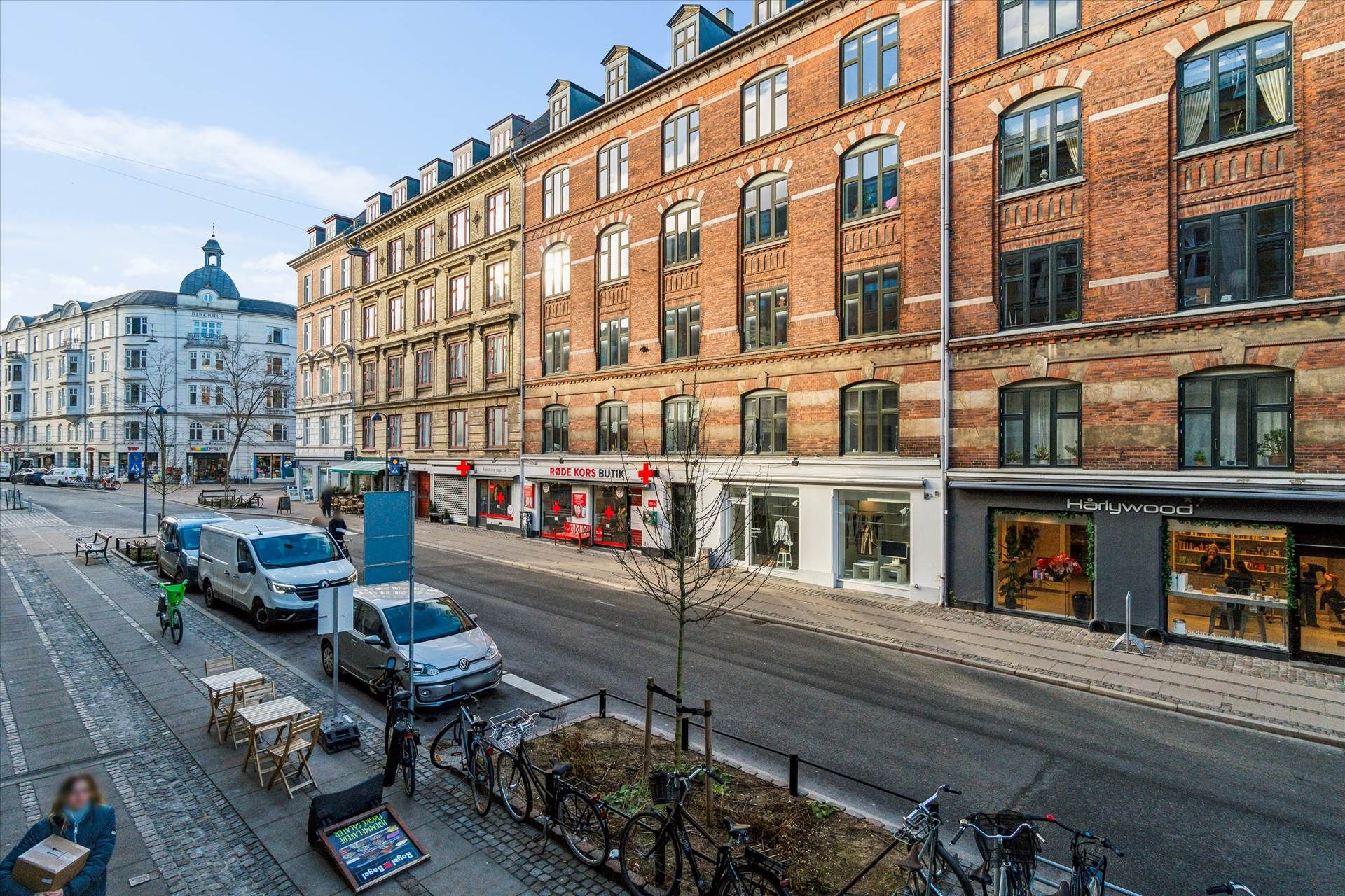 Gade med parkerede biler og cykler, store bygninger og butikker med udsalgsvinduer, træer og bordsætninger langs fortovet