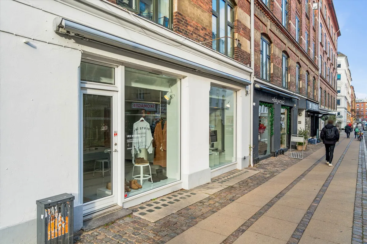 Butiksfronter med store ruder og udstillinger af tøj og sko i et moderne byområde med travle fodgængere og grønne potter