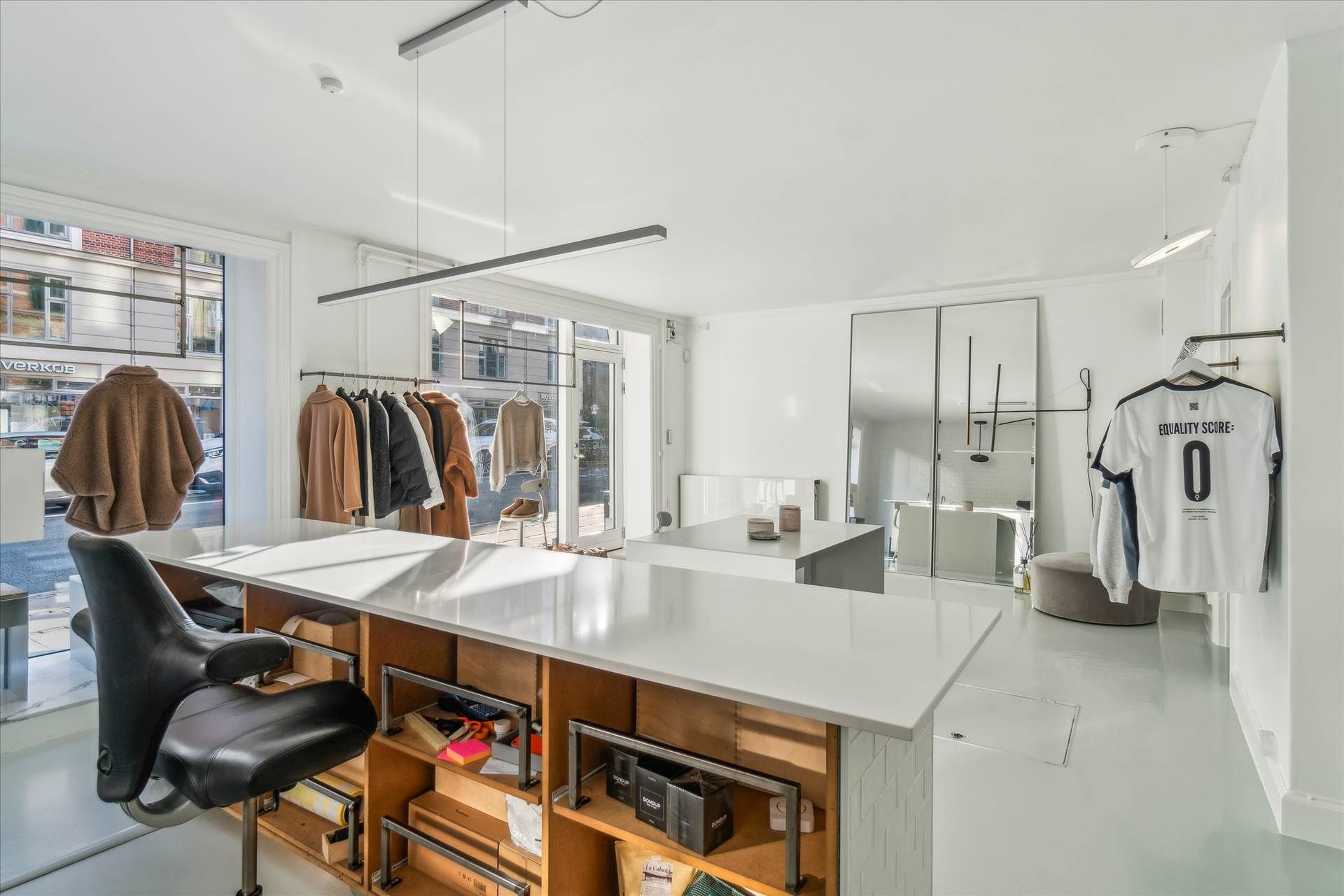 Inde i en forretning er der en hvid væg og gulv, tøj på ophæng, et bord med sæde, et spejl og en trøje med nummeret 0