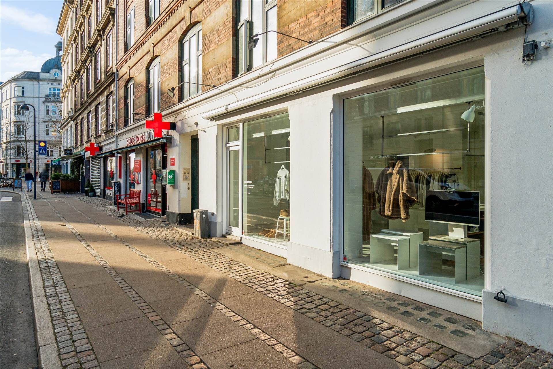 Erhvervslokaler med glasfront og rødt kors i København, hvor butikkerne er åbne og folk går forbi.