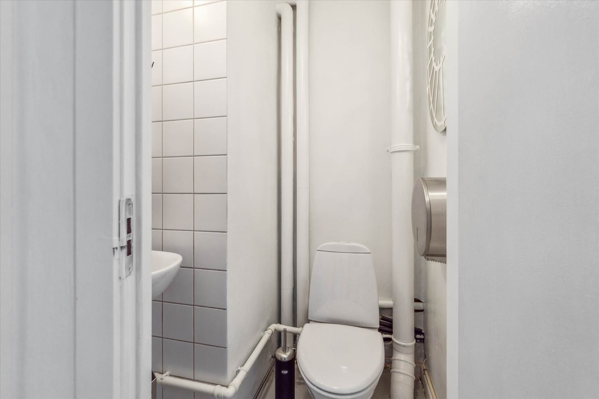 En lille, hvidt dekoreret toiletrum med toilet, håndvask og to toiletpapirholdere, samt hvide, horisontale fliser på væggen