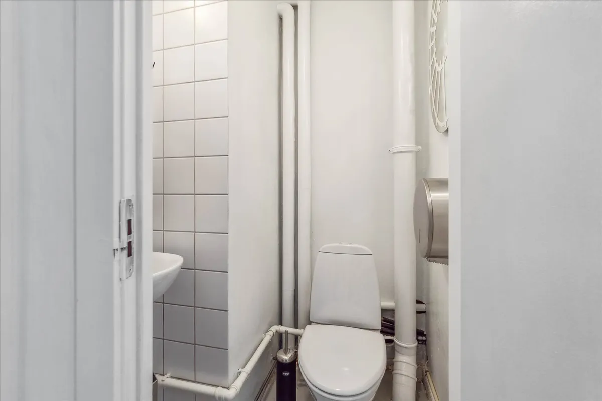 En lille, hvidt dekoreret toiletrum med toilet, håndvask og to toiletpapirholdere, samt hvide, horisontale fliser på væggen