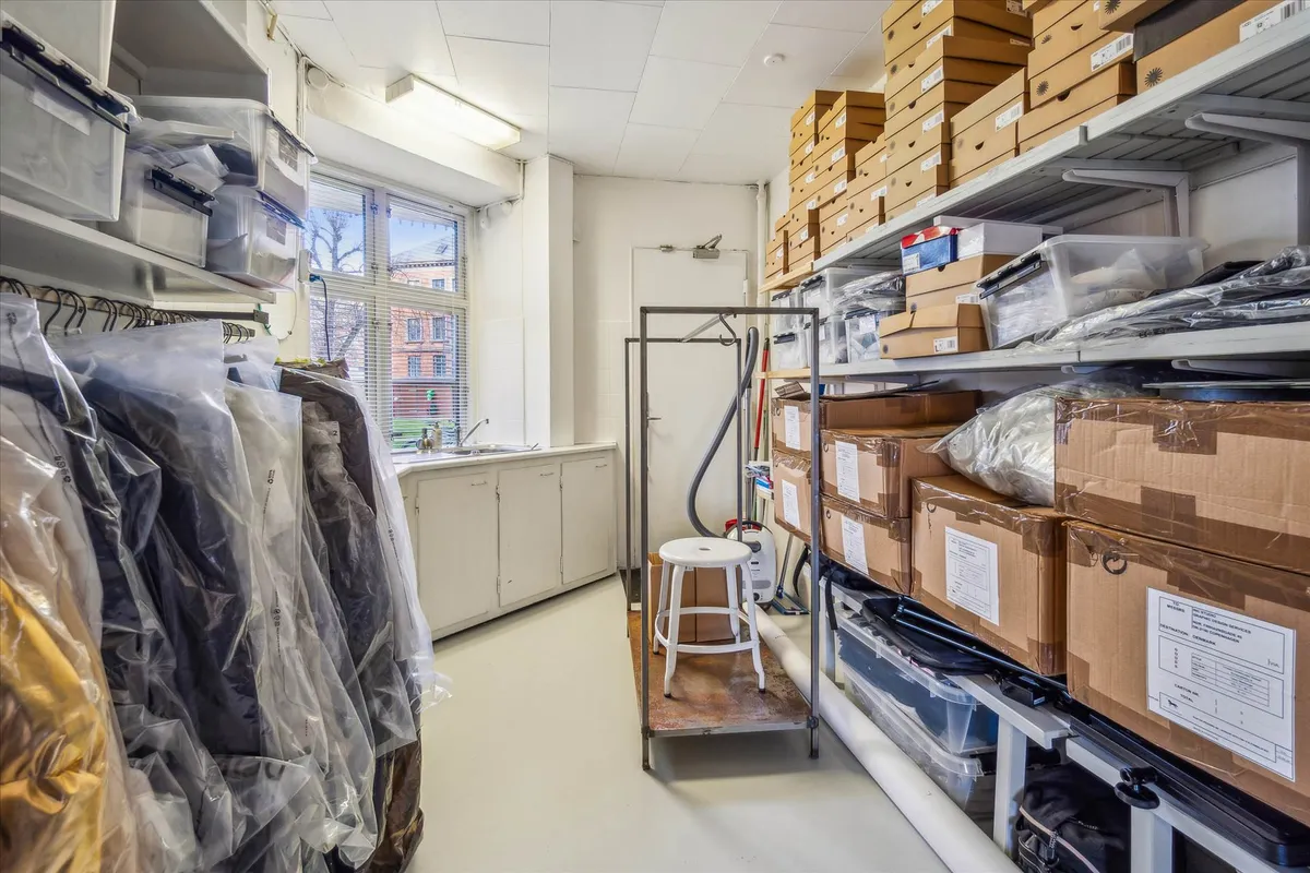 Lagerrum med plastasker, emballagemateriale, kartoner og stol samt tøjvask og skraber i baggrunden