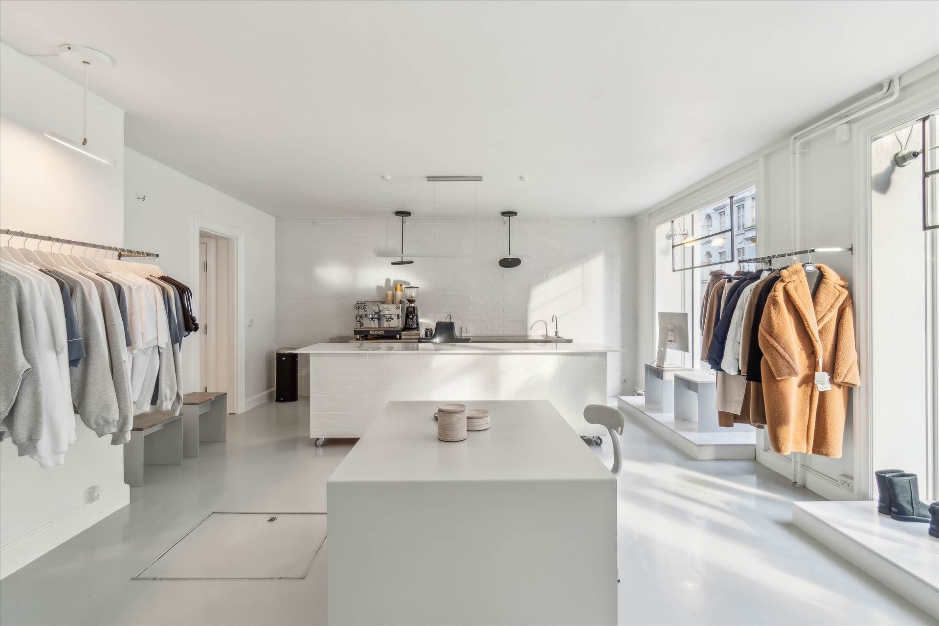 Hvidt interiør med lysegul gulvbelægning, hængende tøj og møbler, lysekroner, køkken med kaffemaskine og vask samt