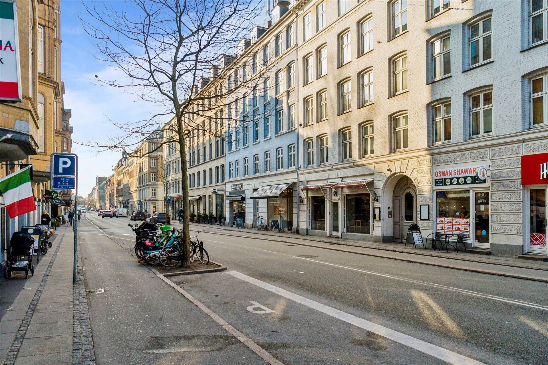 Vej med butikker og huse på Frederiksberg C. En italiensk flag, parkerede cykler og restauranter langs vejen. Solskin og