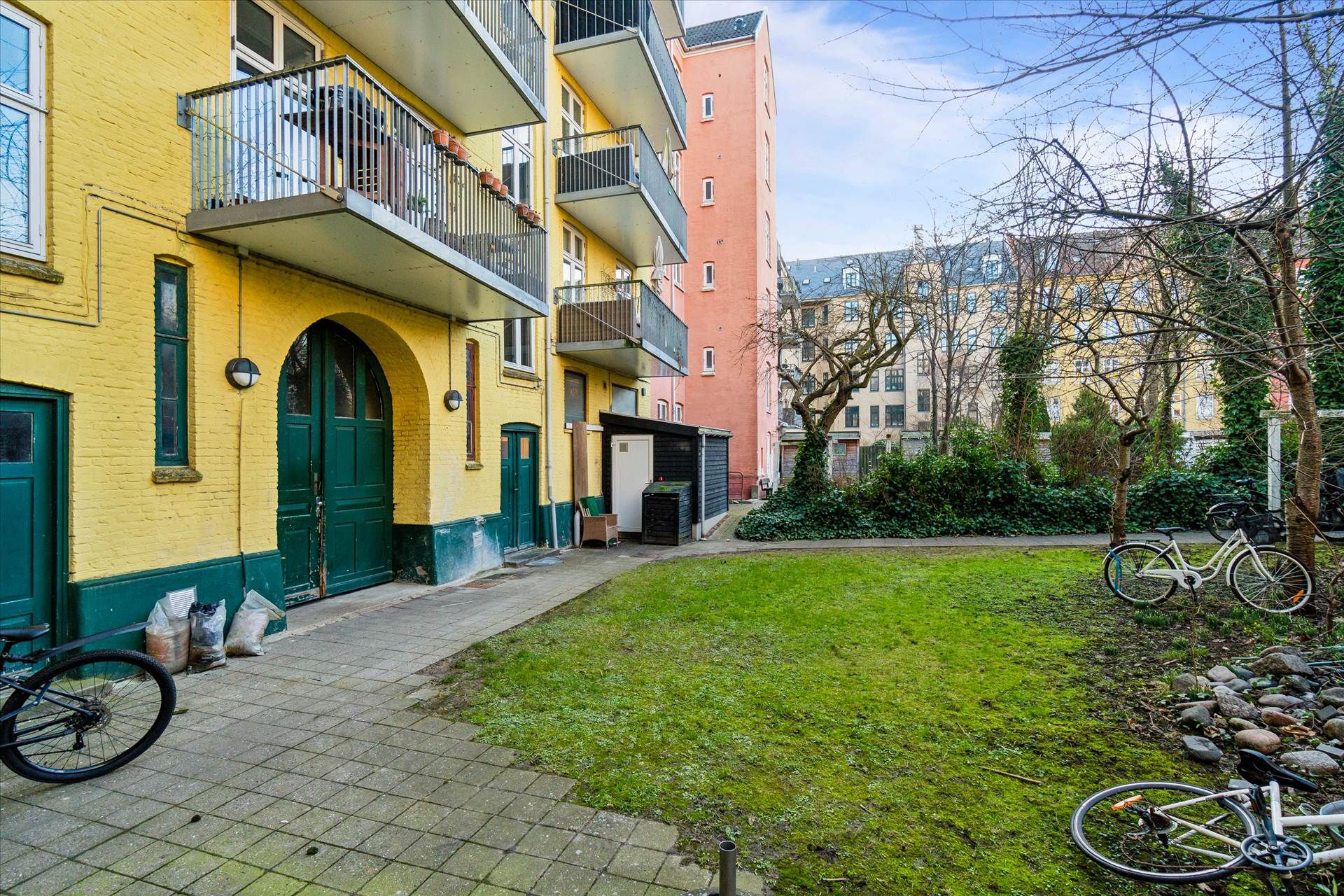 En grøn- og gulbygning med balkoner og to grønne døre samt to cykler parkeret i baghaven med mosede sten.