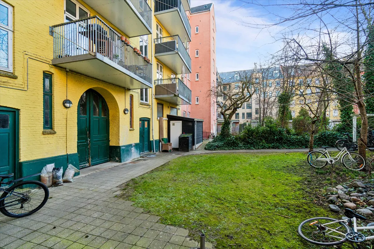 En grøn- og gulbygning med balkoner og to grønne døre samt to cykler parkeret i baghaven med mosede sten.