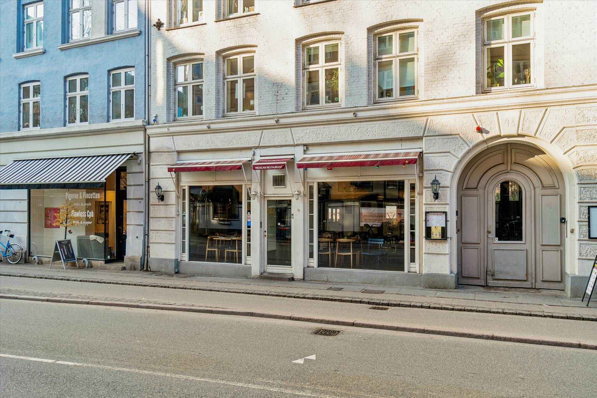 Forretning med flere vinduer og indgangsdør med bueform og navn. Højre for restauranten er en cykel parkeret.