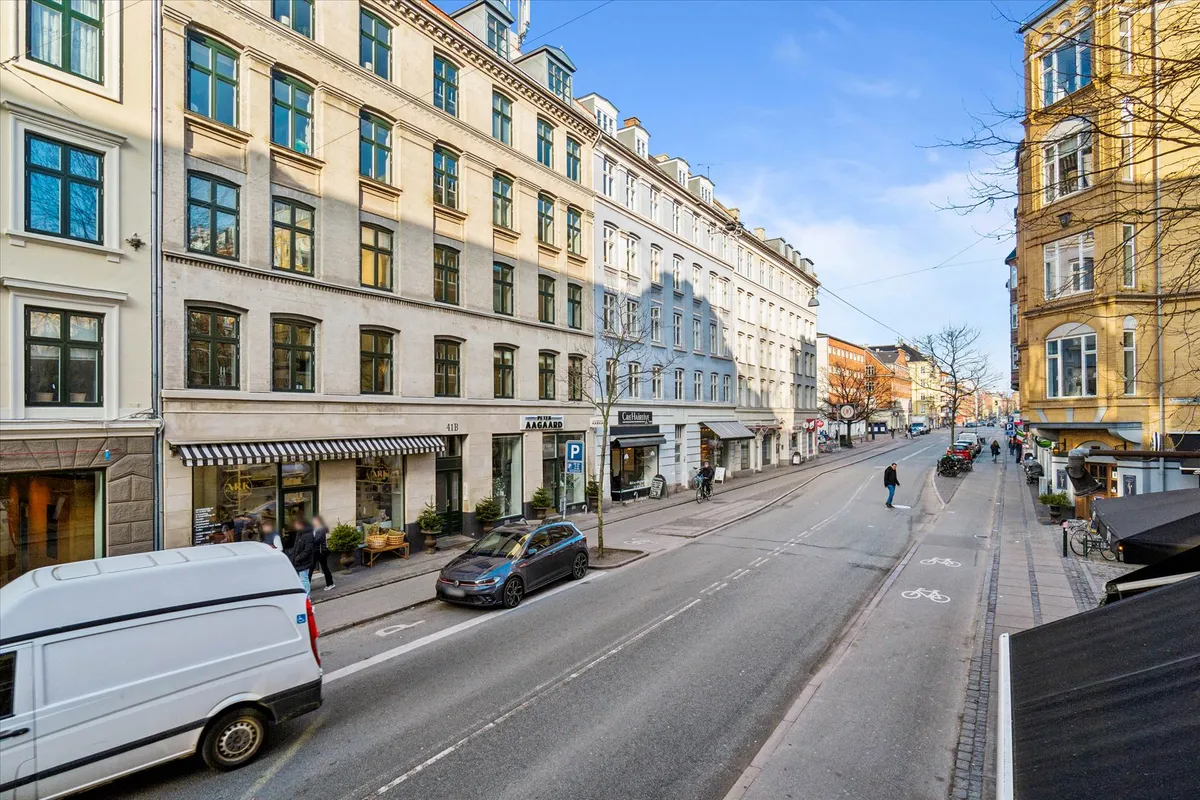Moderne bygning med mange vinduerne og to biler parkeret udenfor, samt mennesker og cykler langs gaden