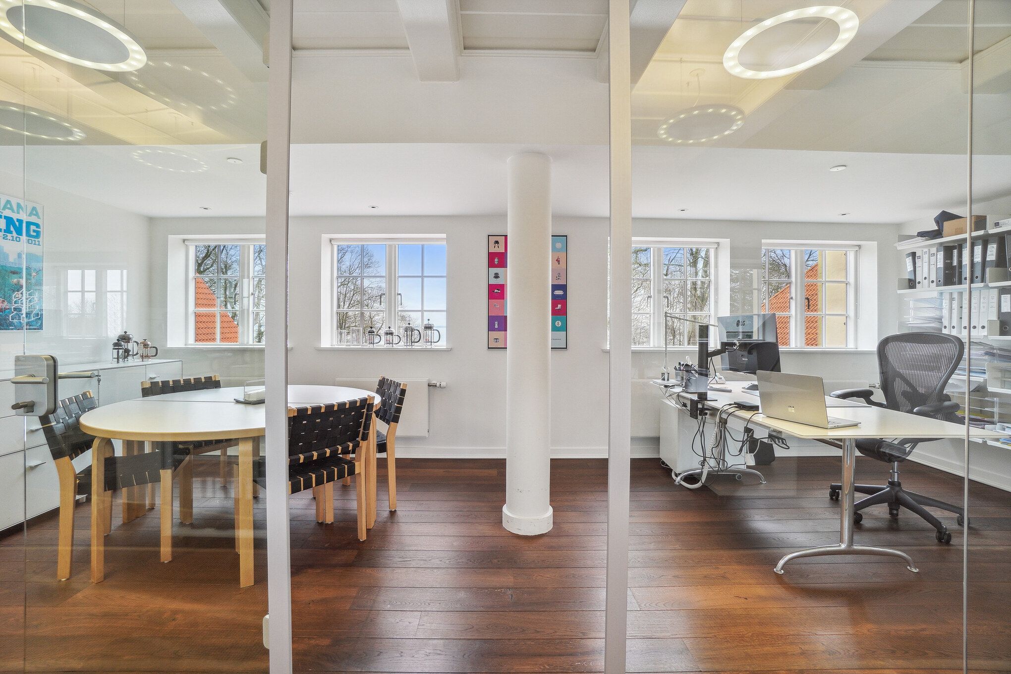 Kontor med moderne interiør og lyse gulve, flere vinduer og lyse runde loftskroner. Rundt bord med stole og en skrivebord