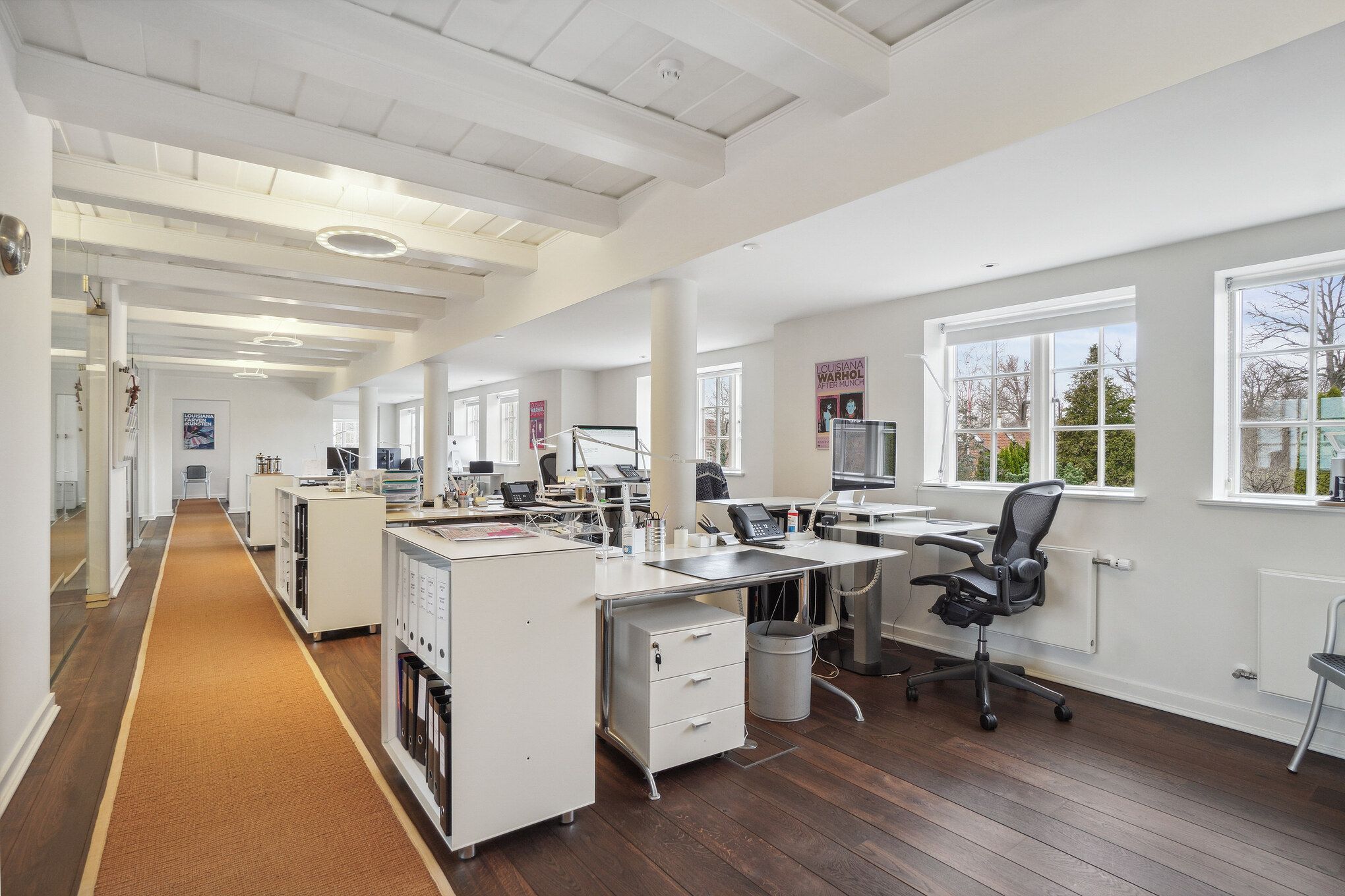 Kontor med hvide vægge og gulv, lyse beige loftsbjælker, moderne møbler, computere, stole, skraldespande, lysekroner og ruder