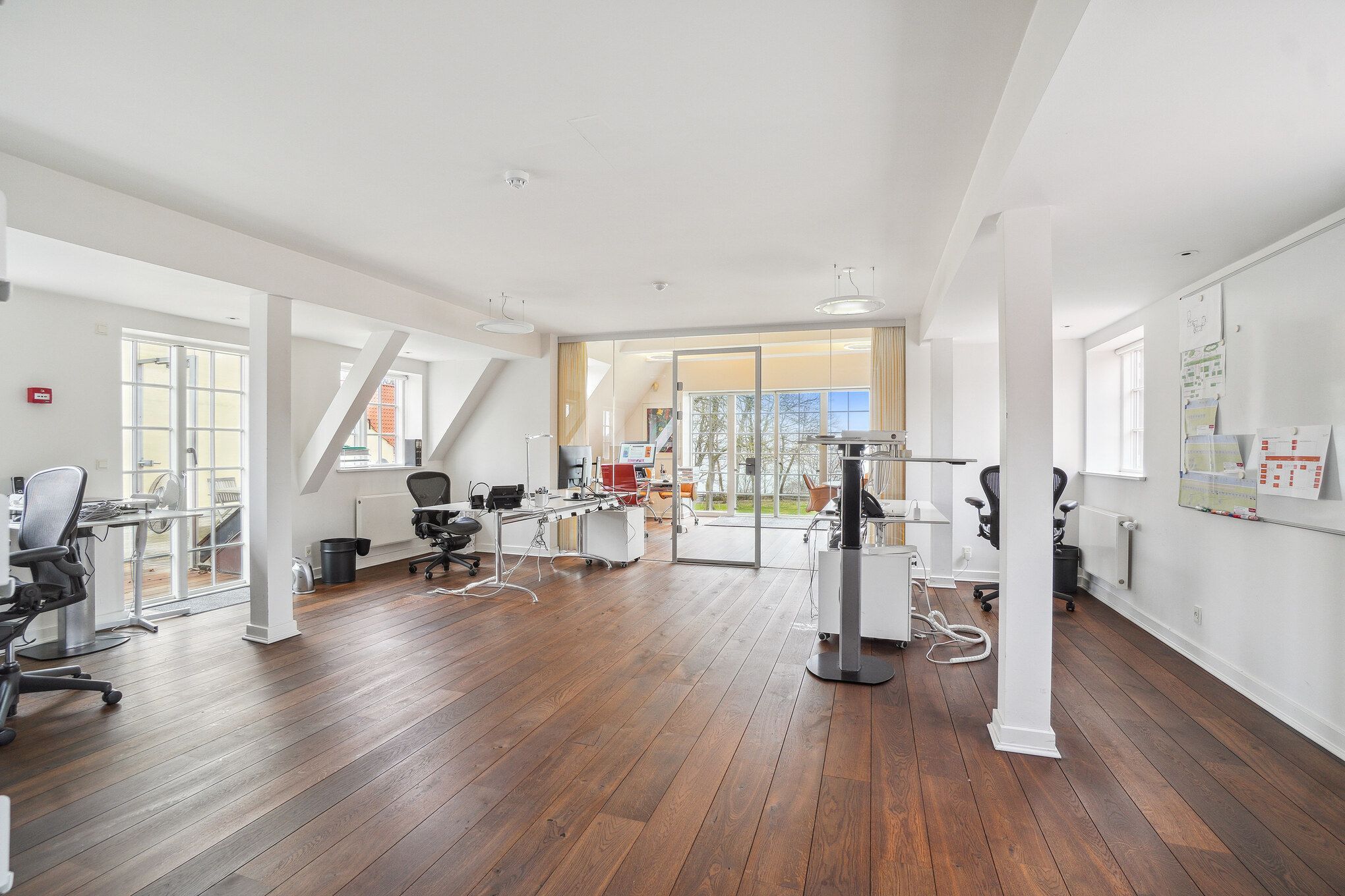Kontor med højloft og møbler i lyse farver, højt teknologisk udstyr og vinduer med udsigt til skov og bygninger