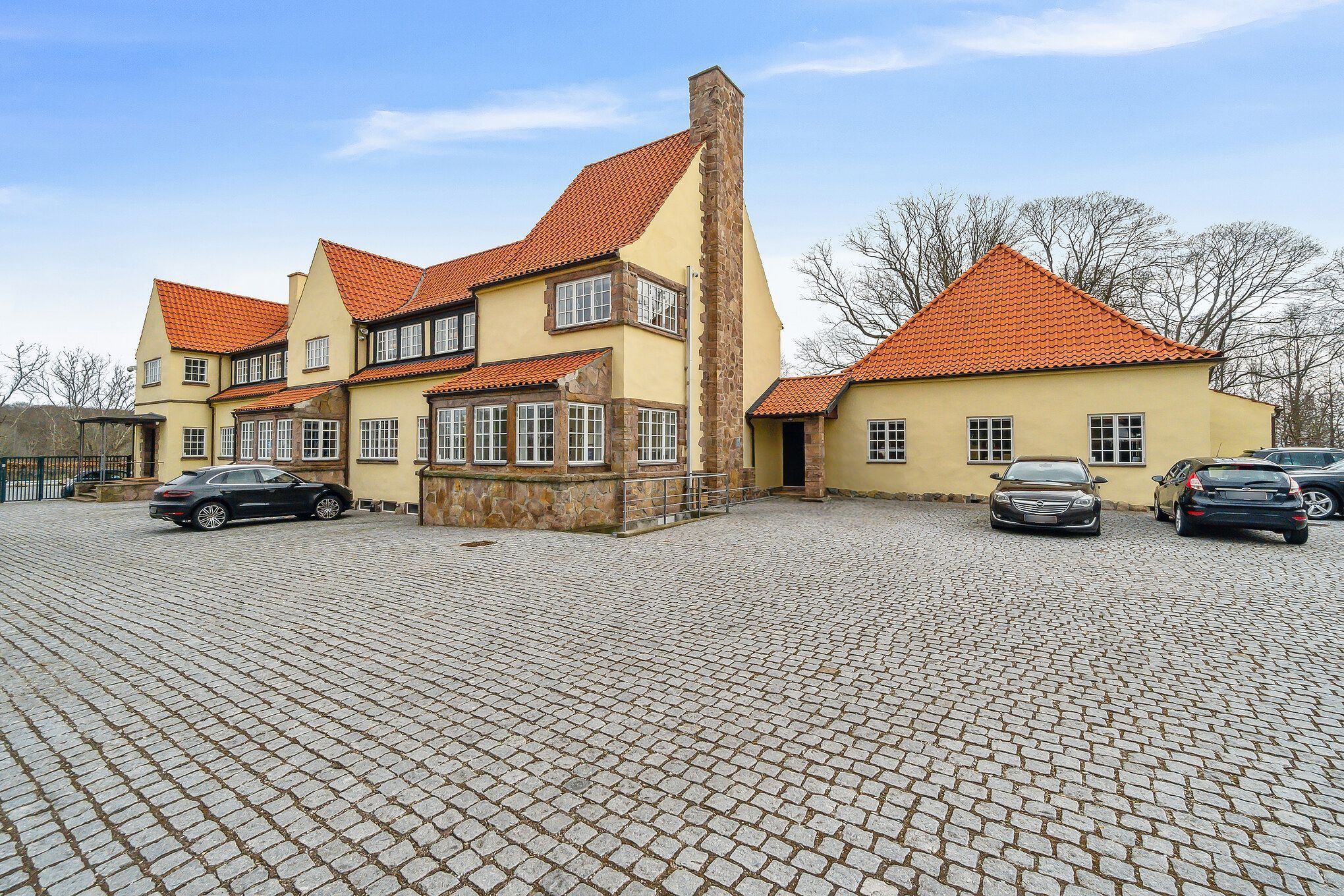 Moderne og stilfuld villa med to separate bygninger, hver med rødt tag og hjørnesten. Foran er parkeringsplads med flere