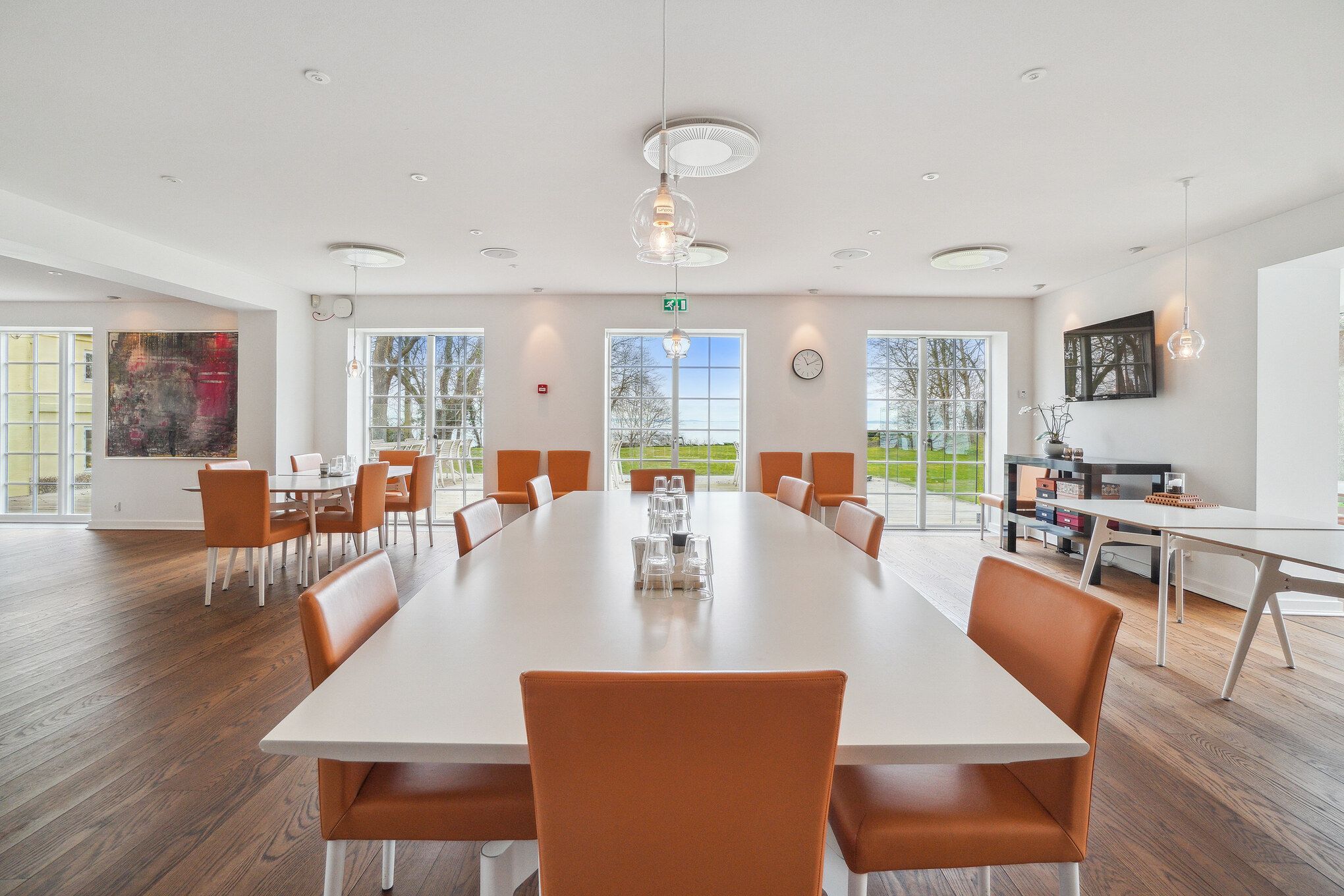 Luksuriøs konferencesal med lyse trægulve, hvide borde, orange stole, store glasvinduer og udsigt til grønne marker