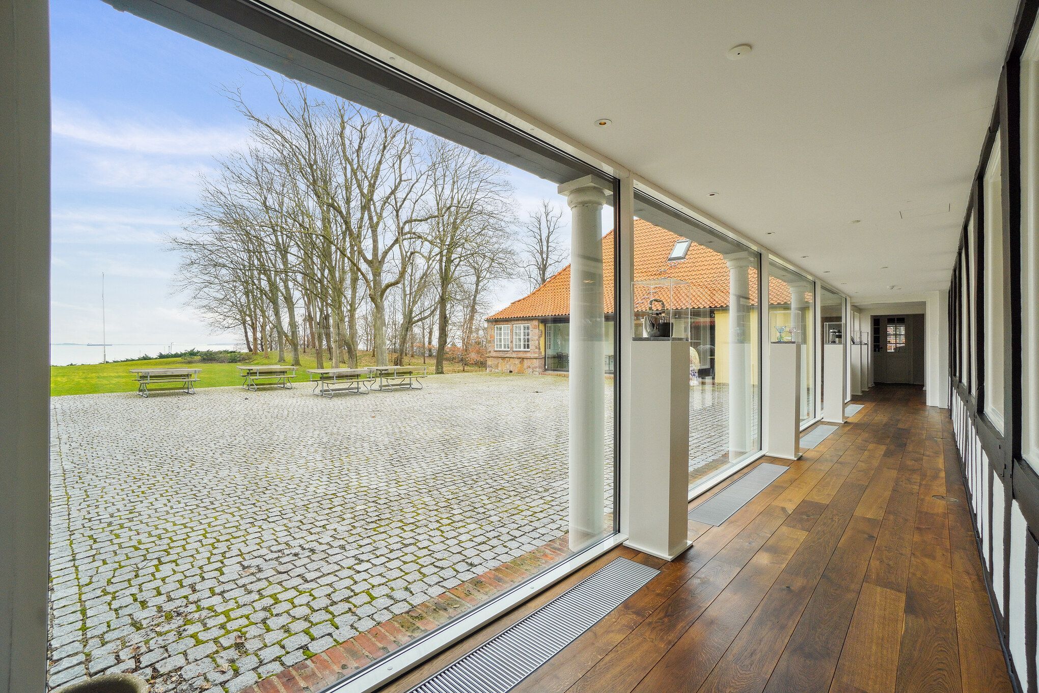 Elegant, moderne korridor med glasvinduer og højloft. Trægulv og hvide vægge. Udsigt til have og bygning med tegltag og