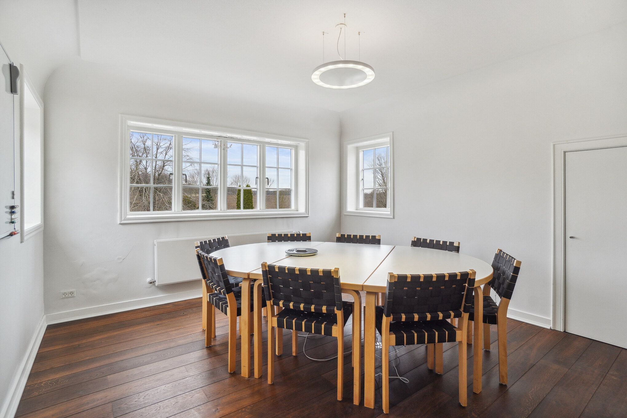 Moderne mødelokale med højt loft og rundt, hvidt loftslys, runde borde med lyse stole, og 3 store vinduer med udsigt til