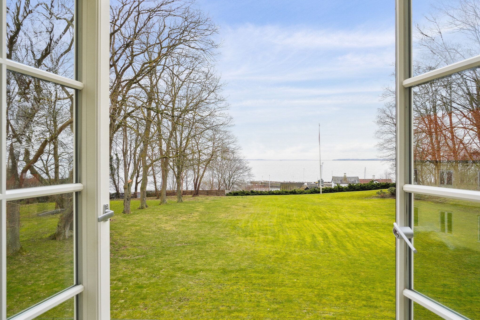Vindue med udsigt til grøn marker og træer med udsigt til havn og bygninger i baggrunden