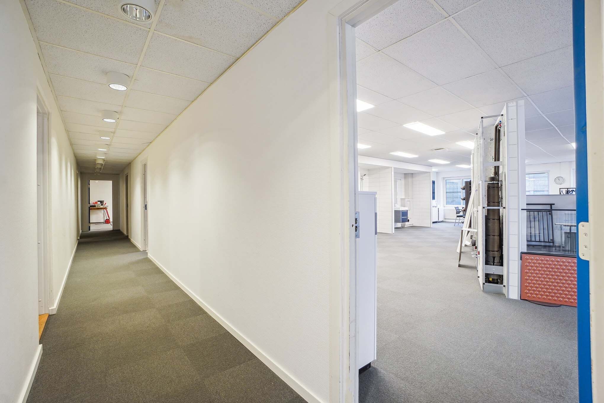 Lang og tom gang med hvide vægge, lysekroner, gulvbelægning og åbne døre til kontorlokaler med møbler og udstyr