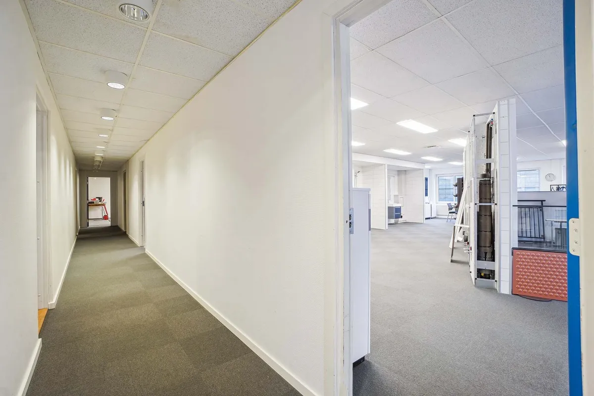 Lang og tom gang med hvide vægge, lysekroner, gulvbelægning og åbne døre til kontorlokaler med møbler og udstyr