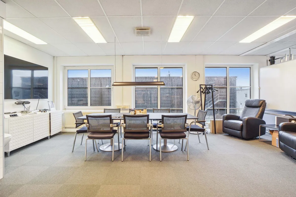 Kontorrum med lyse gulve og store vinduer, rekreationsområde med lænestol og sofa, skrivebord med flere stole og tv og en