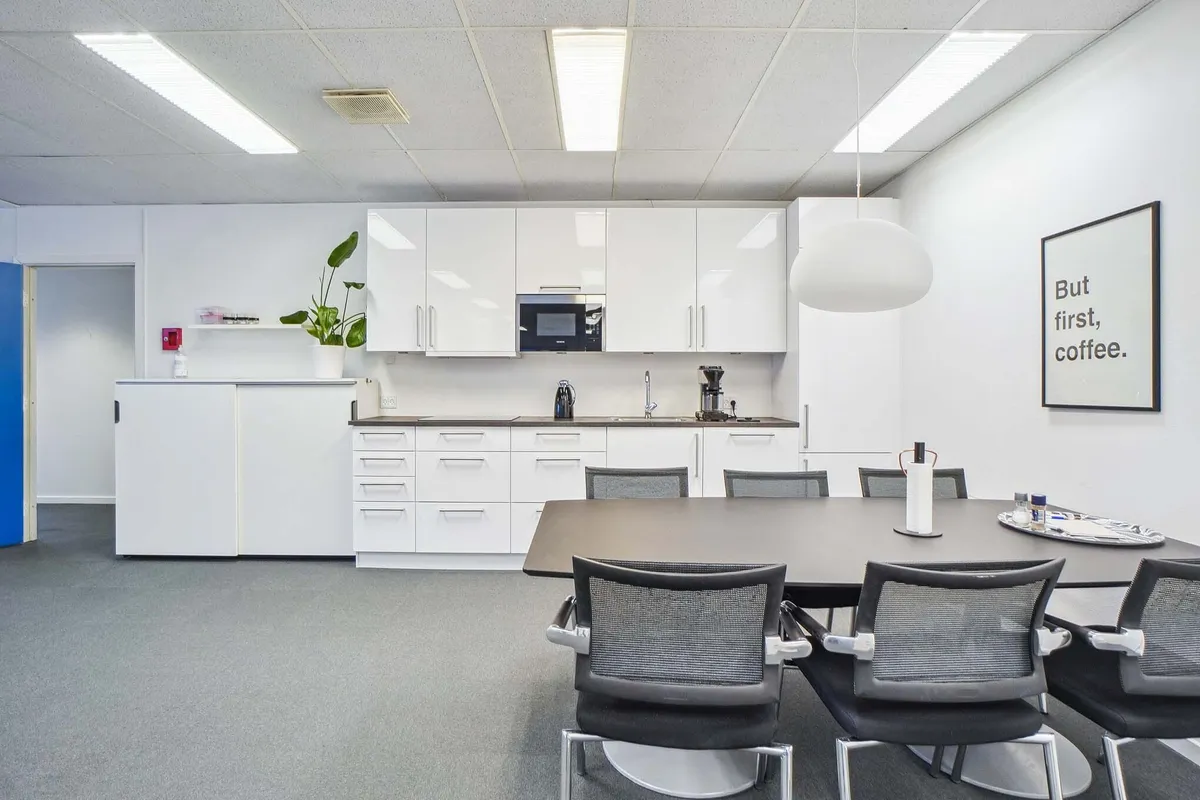 Moderne mødelokale med hvide vægge og hvide skabe, hvor der er en køkkenhjørne med mikroovn og kaffemaskine samt et bord med
