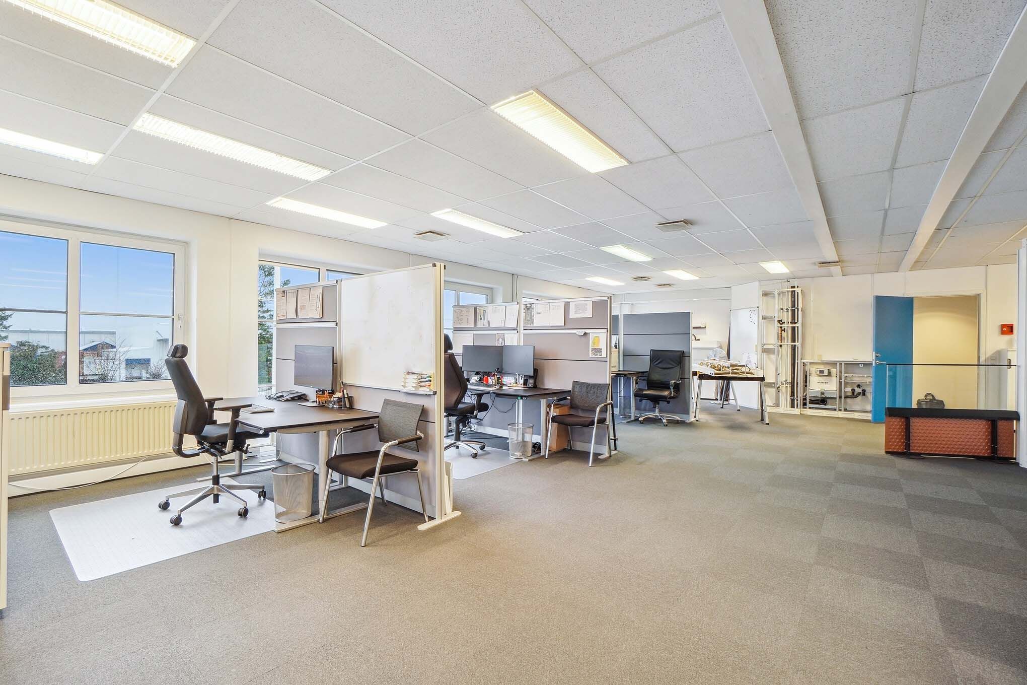 Kontor med lysegult loft, højt vinduesparti, lysegrå gulvbelægning og flere arbejdsborde med computere, stole og kabinet.