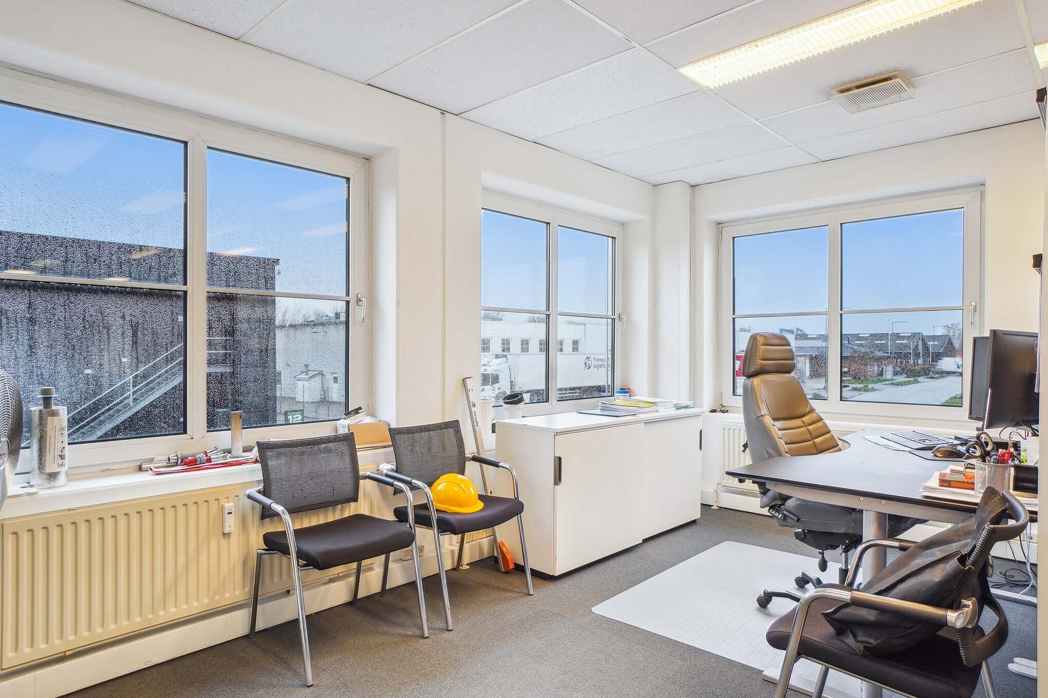 Kontor med tre stole og en skrivebord med flere bøger og en computer. I vinduet står der en hårdhat og værktøj. Der er også