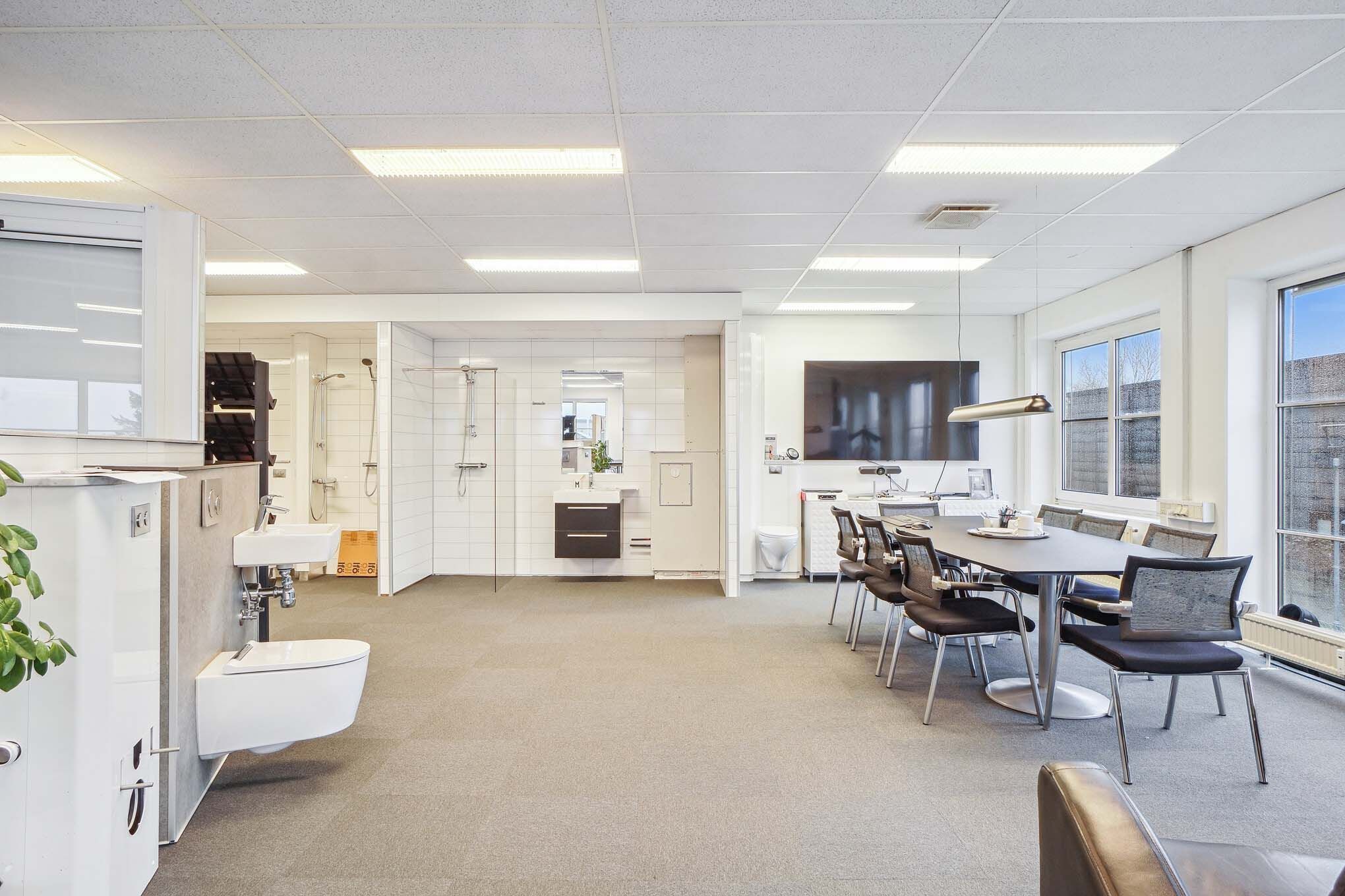 Indenfor er der en moderne kontorlounge med en stor rund borde, stolpe og tv, og i baggrunden er der en hvid badeværelse med