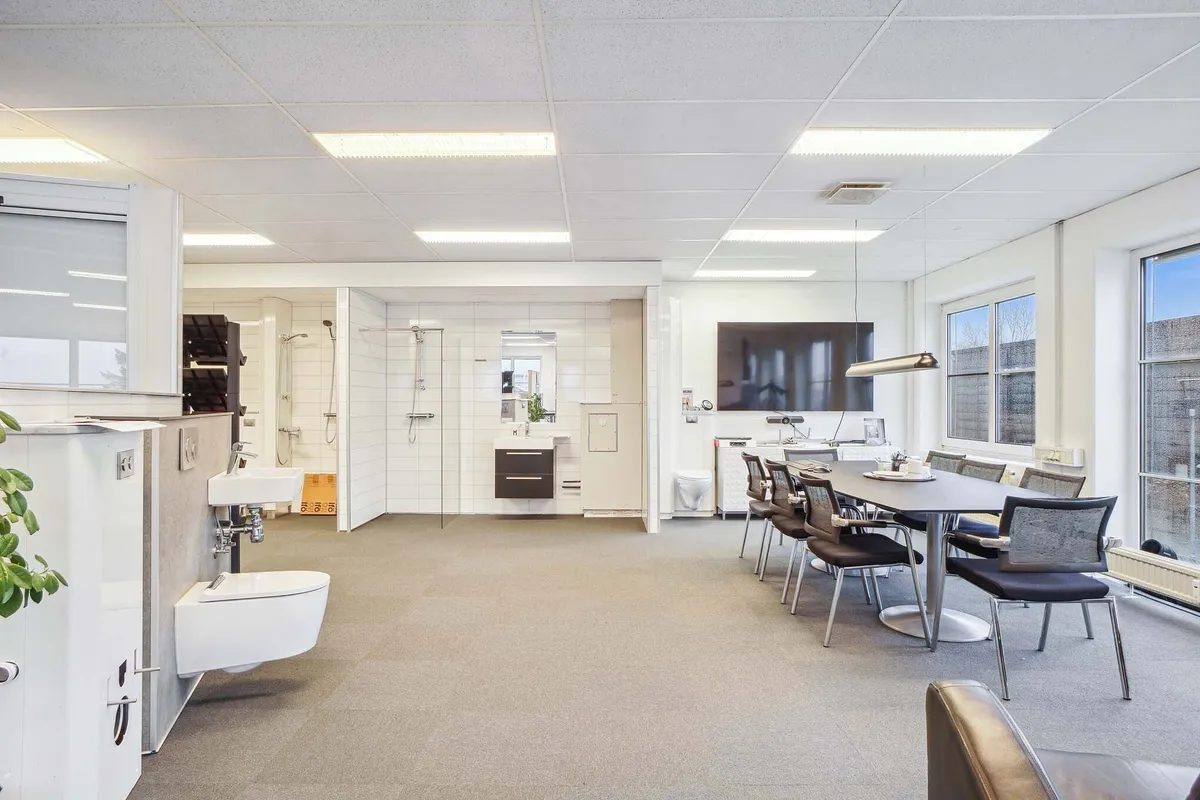 Indenfor er der en moderne kontorlounge med en stor rund borde, stolpe og tv, og i baggrunden er der en hvid badeværelse med