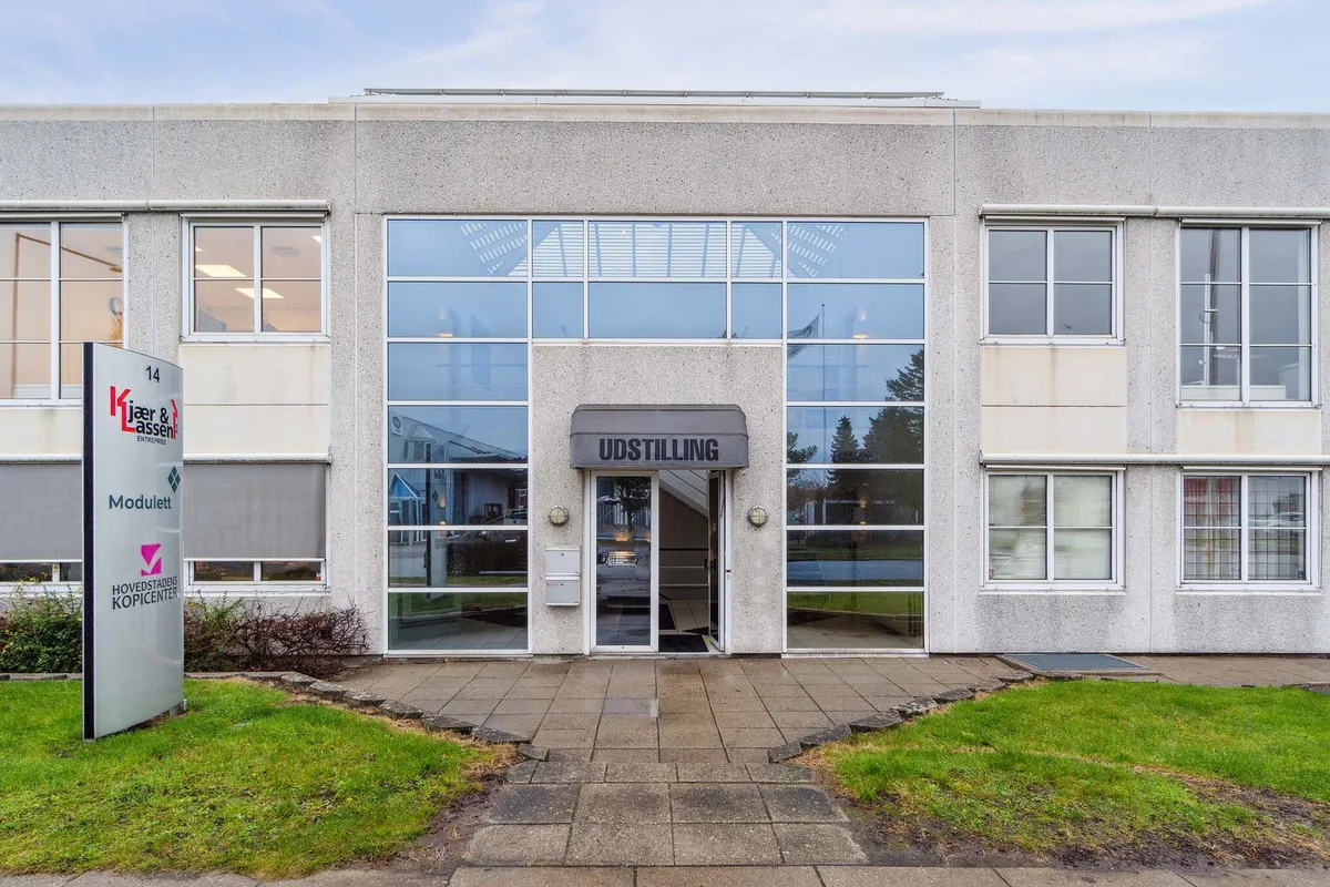 Et bygningsfront med glasdøre og vinduer, stående foran en grøn plæne, med et skilt med logoet Kjaer & Lassen