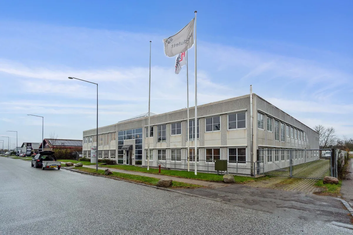 Firmabygning med tre flag og to søjler, indhegnet grund, parkeringsplads og grønne plæner langs vejen med bil og gitterport