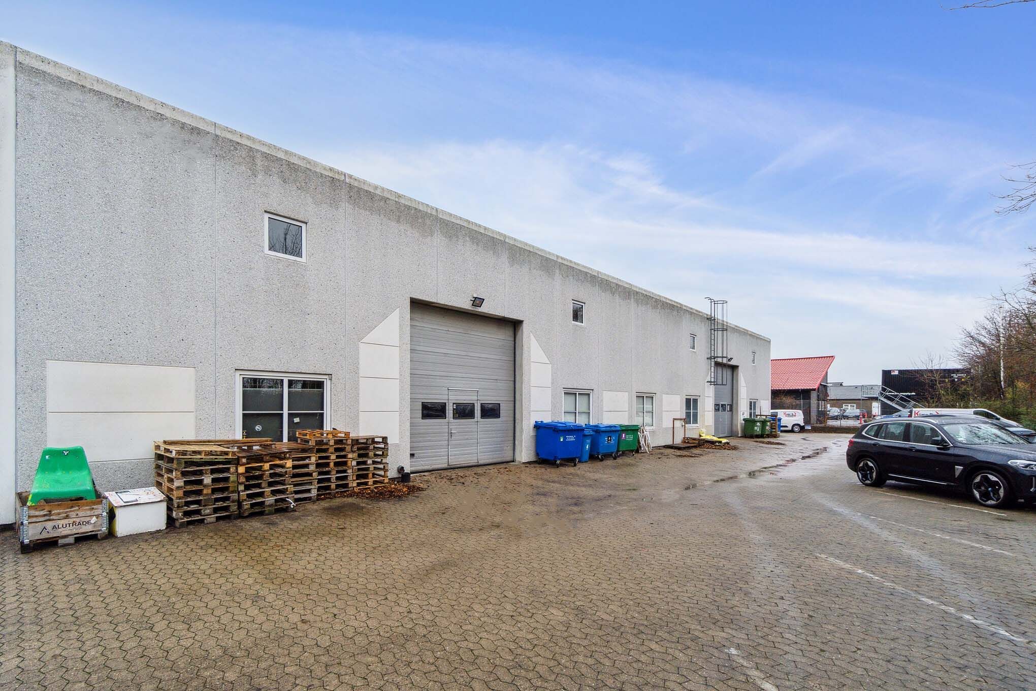 En industriel bygning med garagedøre og flere lagerkasser samt affaldscontainere foran og parkerede biler på siden