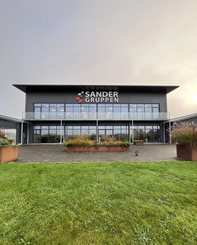 Glasvinduer og to store plantebed i sandfarvede potter uden for Sander Gruppens kontorbygning, der er omgivet af græsplæne