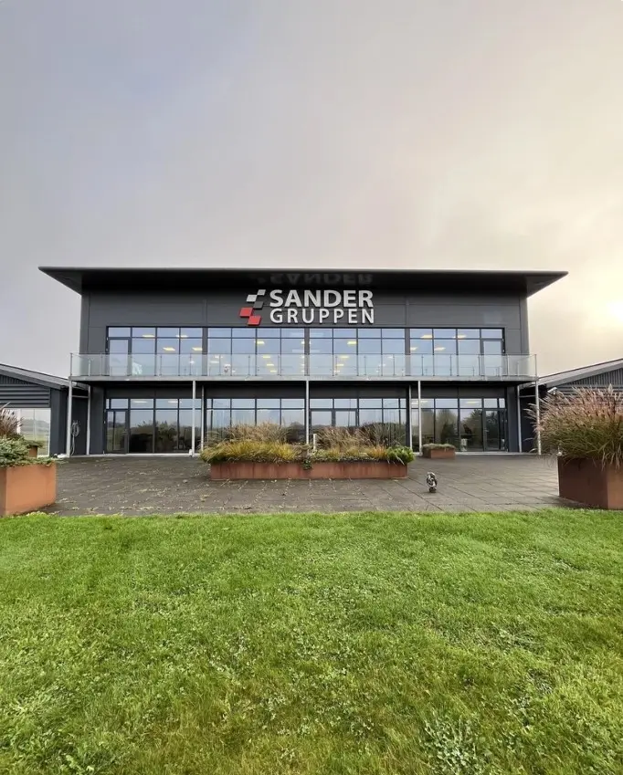 Glasvinduer og to store plantebed i sandfarvede potter uden for Sander Gruppens kontorbygning, der er omgivet af græsplæne