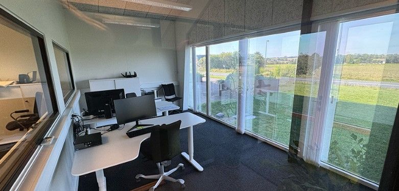 Kontorlokaler med stort vinduesparti med udsigt til grønt landskab og landevej. Kontoret har moderne møbler og skriveborde
