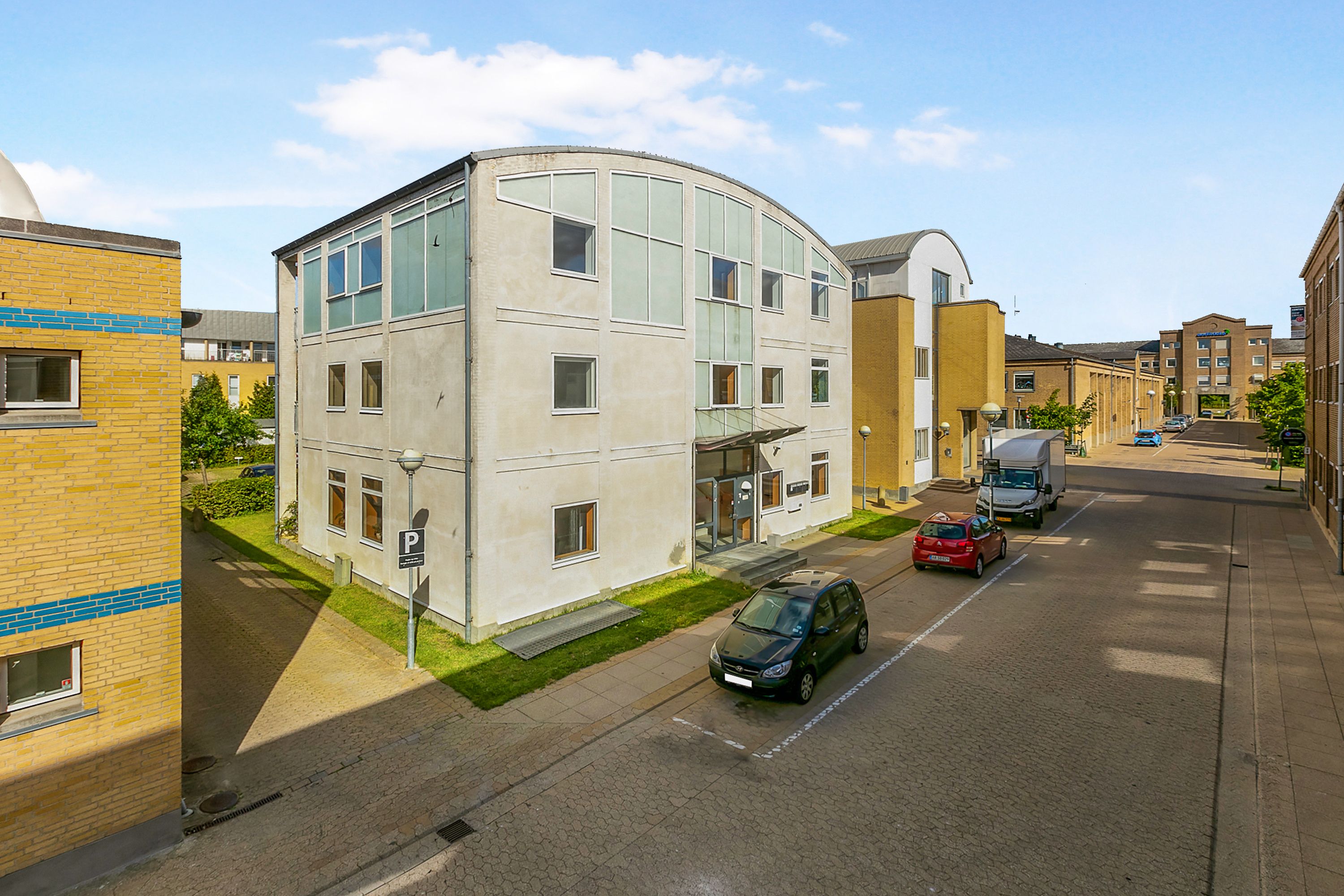 Kontorbyggeri i Taastrup med tre biler parkeret, lyse gulve, en stor grøn plæne og en stor hvid bygning med store vinduer