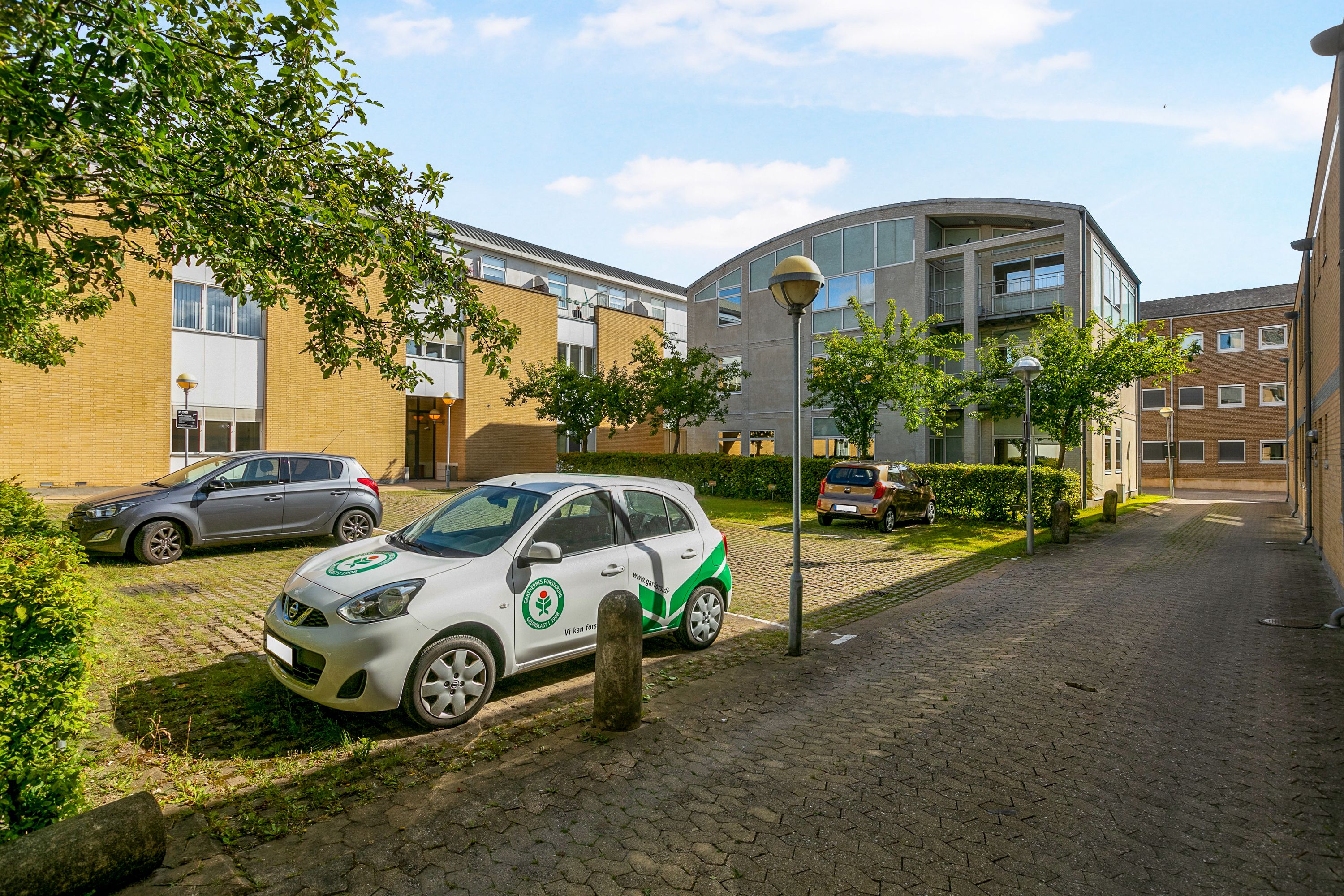 Hvid og grøn bil parkeret på kontorlejlighedsparkering. Moderne bygning med gul stenfasade og glasruder. Buske og træer