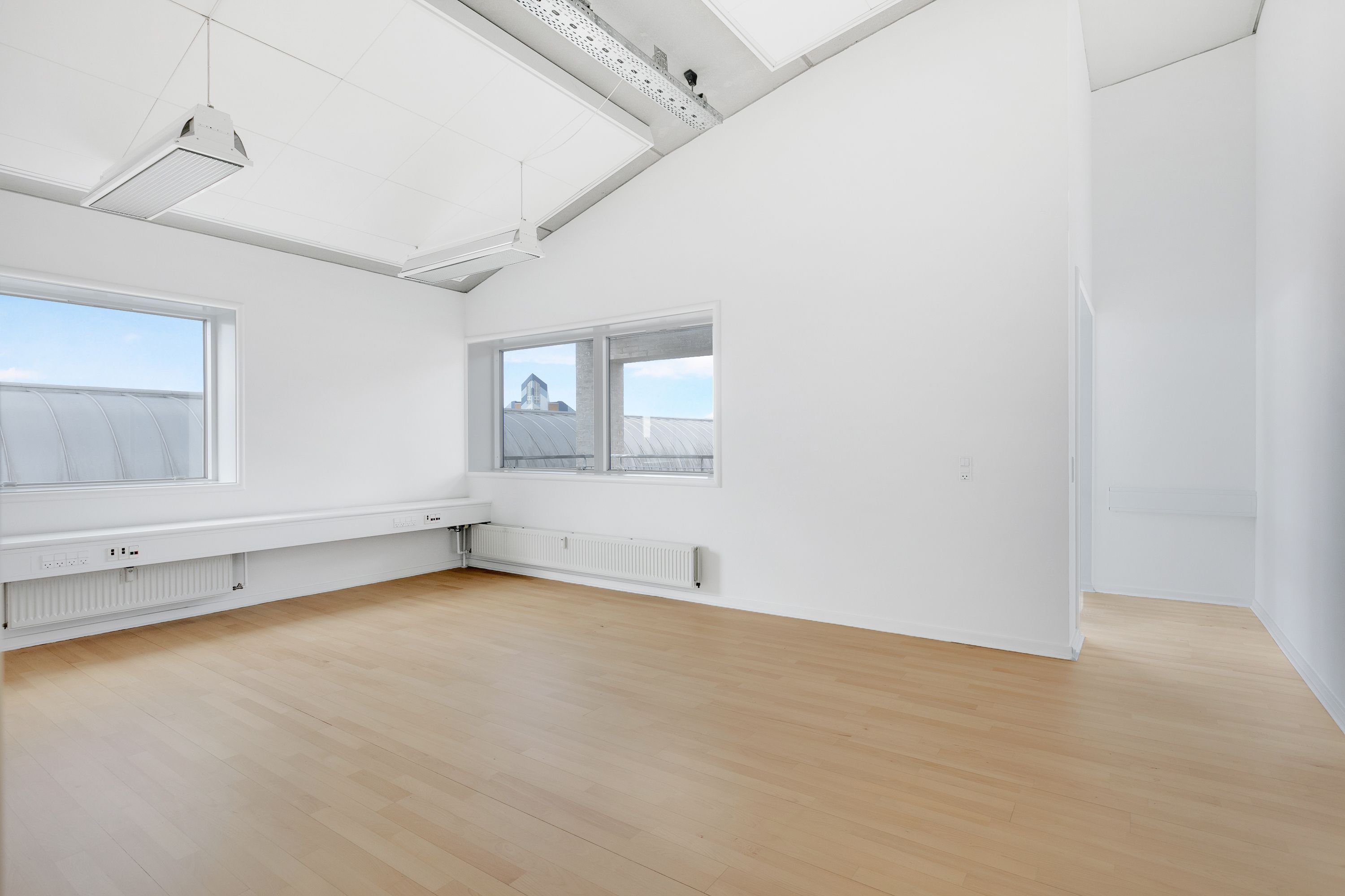 To vinduer med udsigt til byen, højt hvide loft og hvide vægge, lyse og lyse gulvbelægning samt radiatorer under vinduet og