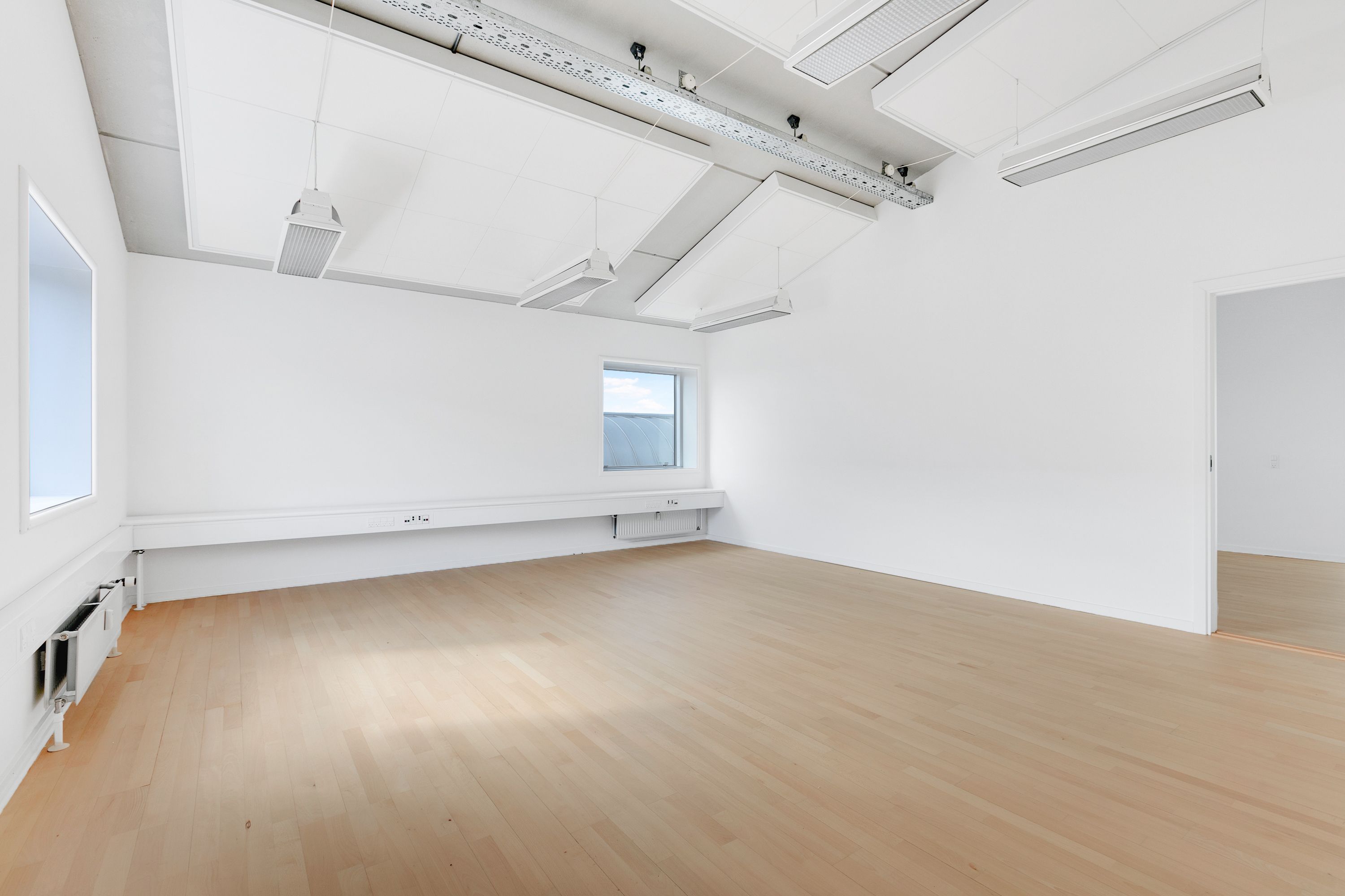 Rummeligt kontorlokal med hvide vægge, lyse trægulve og store vinduer. Der er to radiatorer langs den ene væg og et hvidt