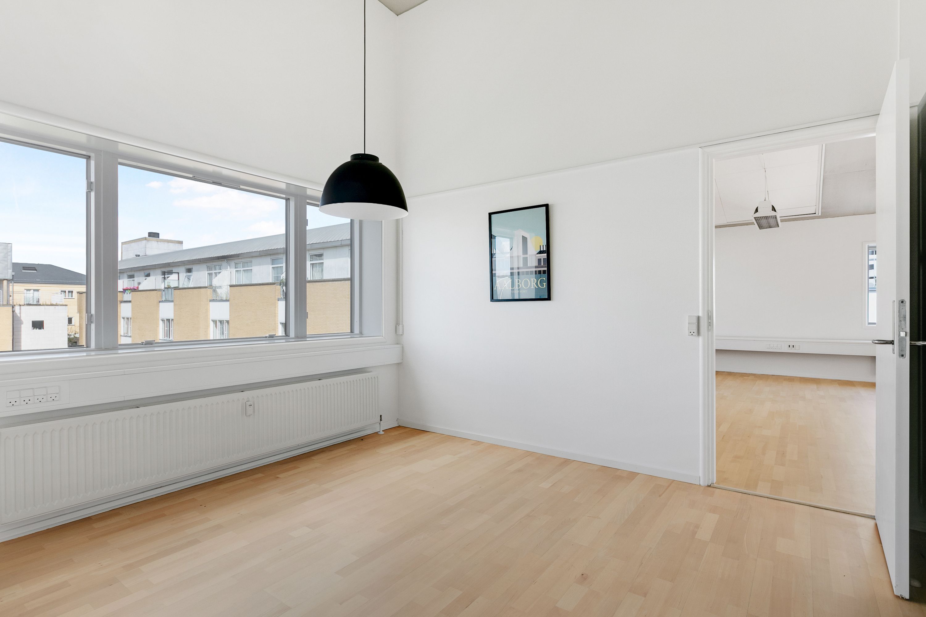 Rummeligt kontorrum med store vinduer og møblerne er fjernet. Væggene er hvide, og der er en åben dør og et kunstværk på den