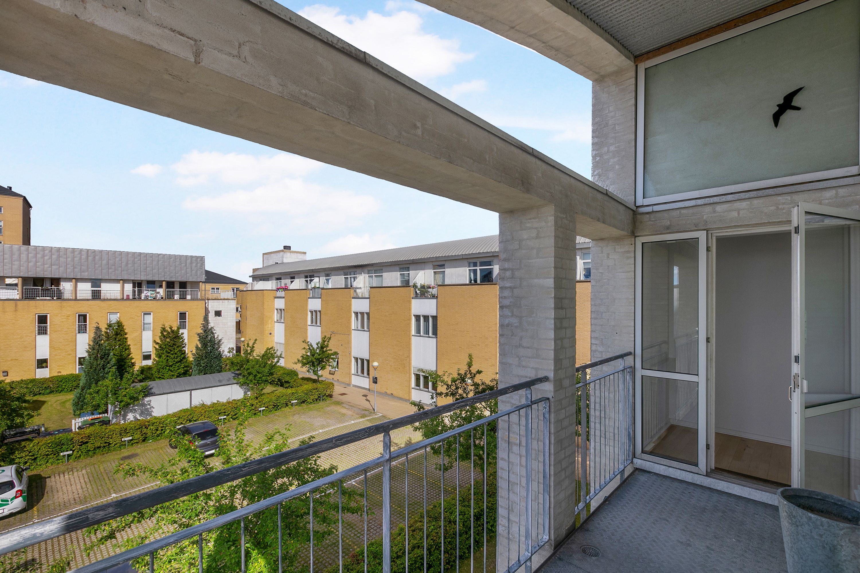 Balkon med metalrammer og glasdøre til lejligheden i multietagers bygning med parkeringsplads og planter i baggrunden