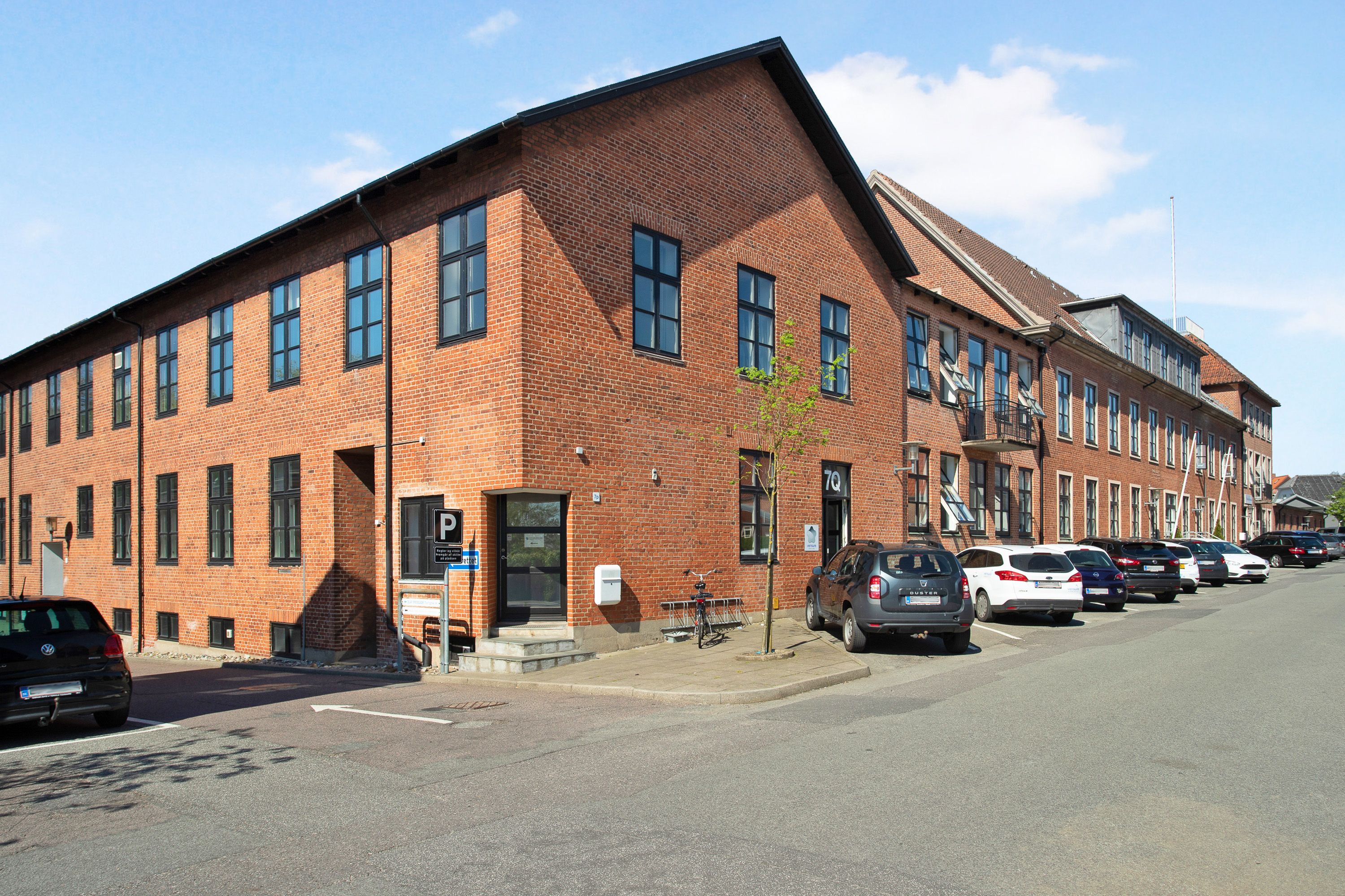 En stor bygning med røde mursten og glasruder parkeret biler foran og et skilt med bogstavet P