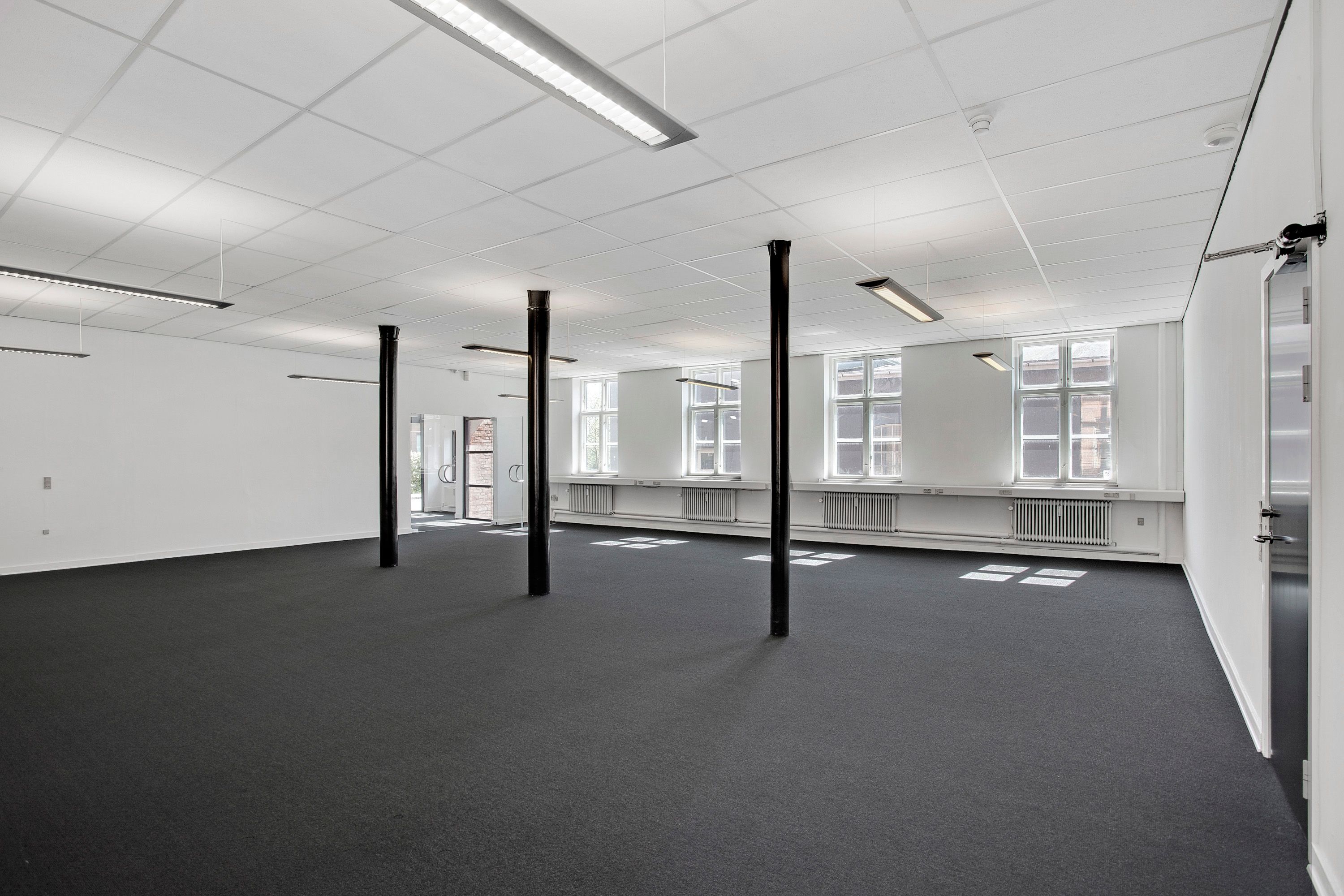En stor, lys, og tom erhvervslejemål med lyse lofter og gulv og flere vinduer og radiatorer