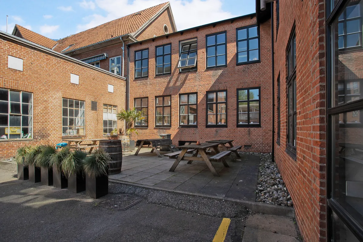 En åben gård med picnicborde, bænke, en stor tønde og plantepotter. Moderne røde mursten og vinduer med dobbeltglas.