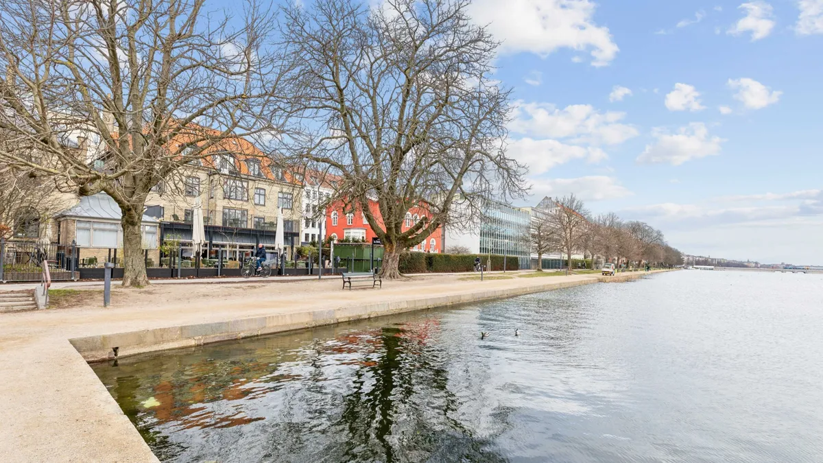 Mennesker cykler langs en park ved vandet og en bygning med orange tag i baggrunden