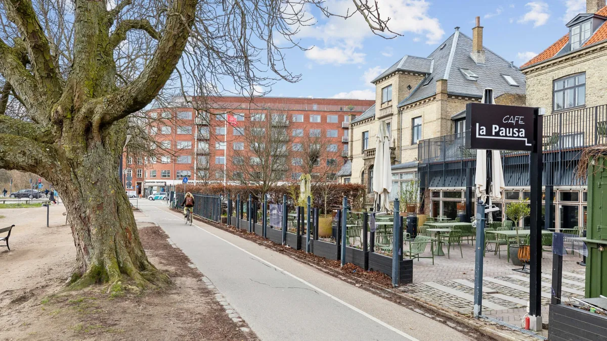 En cykelsti med en stor træstamme og et cafénavn på skriften ved siden af en bygning med terrasse