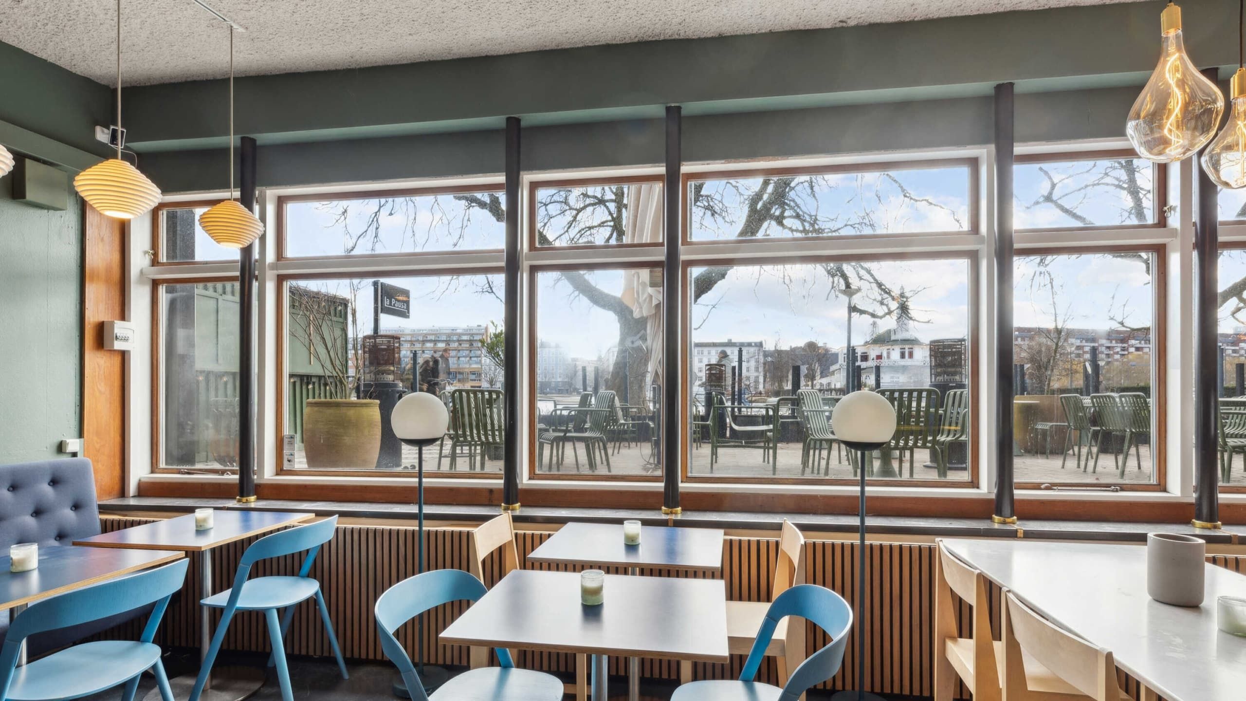 En café med et stort vindue og udsigt til grønne områder og bygninger samt borde og stole med hvide og blå farver