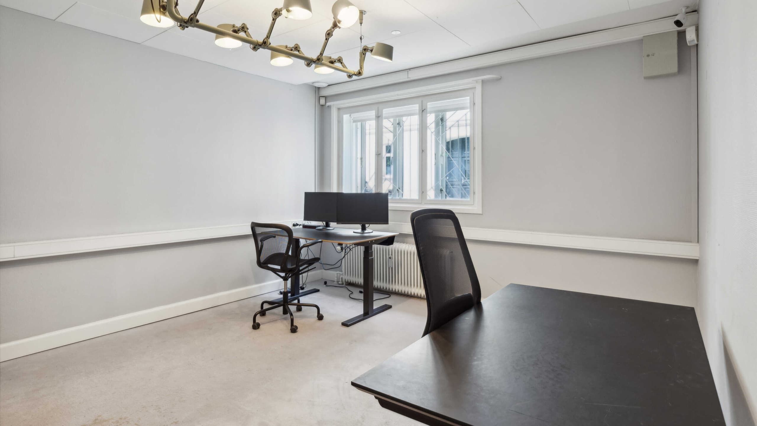 En tom kontorrum med to skriveborde, hver med en computer og en stol. Væggene er hvide og der er en radiator nedenunder