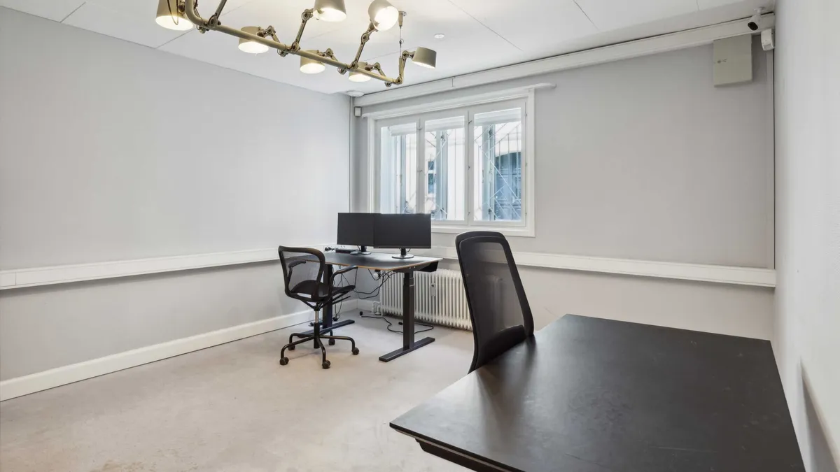 En tom kontorrum med to skriveborde, hver med en computer og en stol. Væggene er hvide og der er en radiator nedenunder